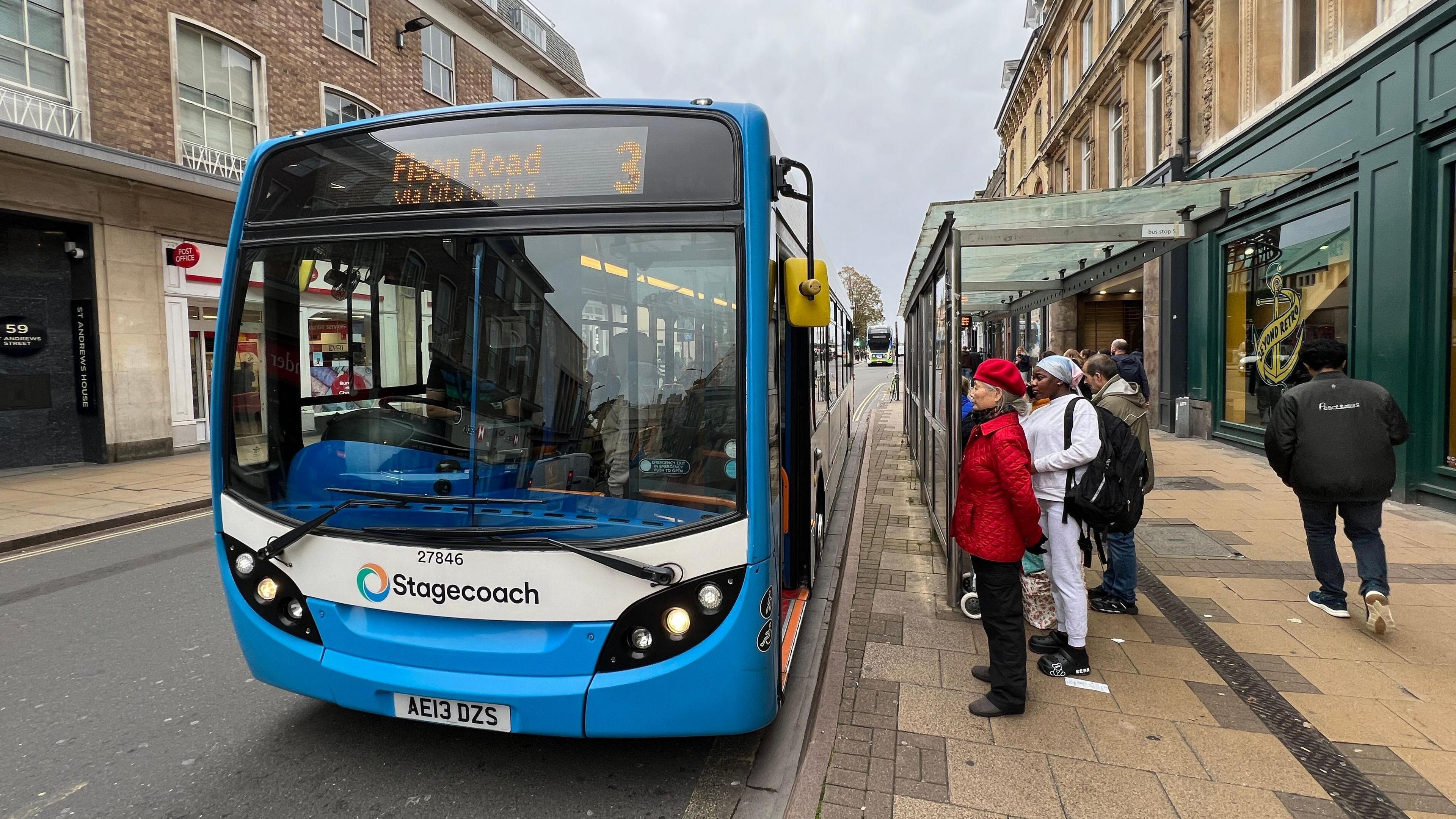 A blue number three Stagecoach bus in a street lined by shops, with passengers waiting to board underneath a bus shelter.