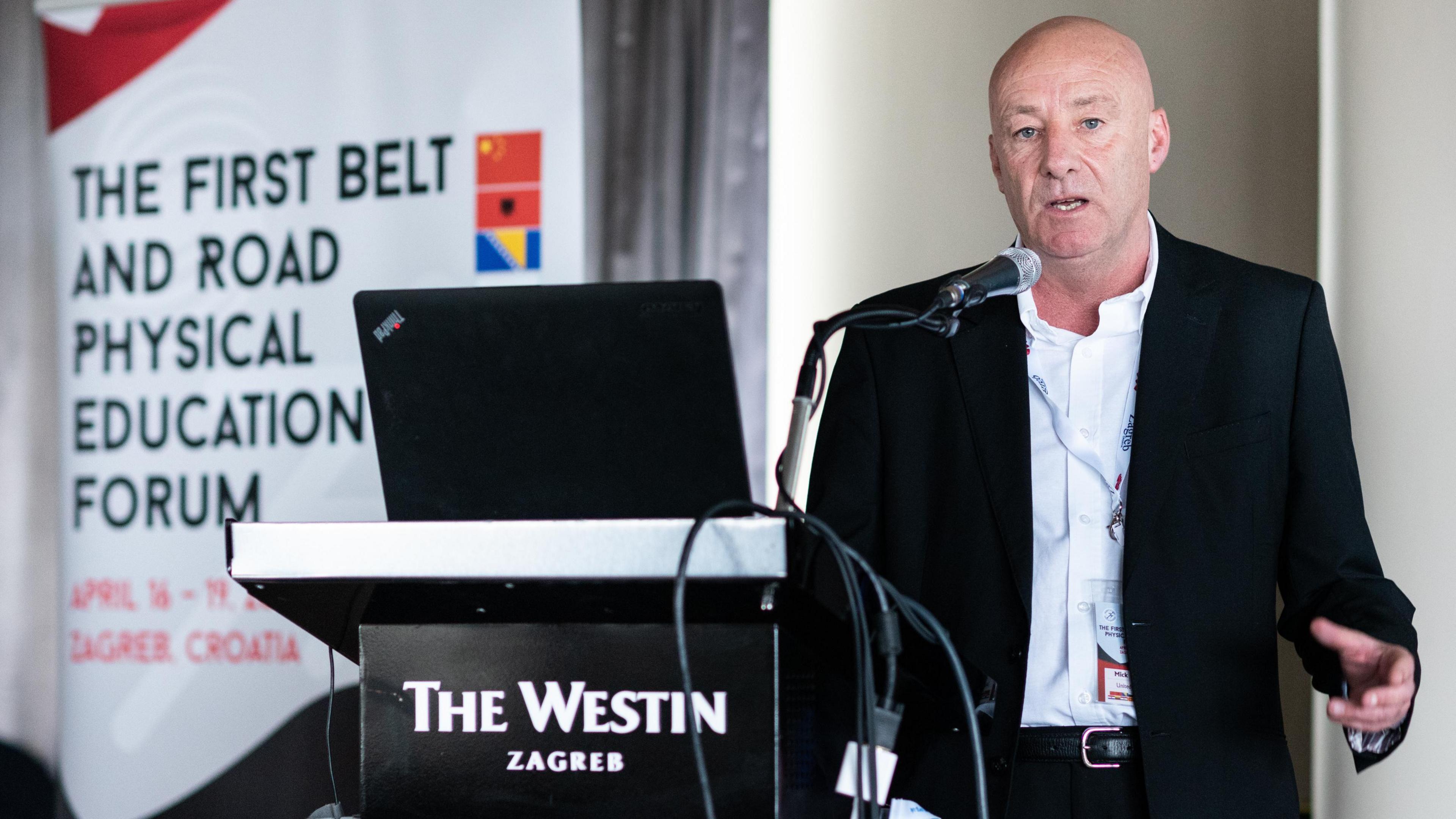 Mick Donovan, a middle-aged man with a shaved head, speaks into a microphone at a lectern labelled "The Westin Zagreb". A banner behind him reads "THE FIRST BELT AND ROAD PHYSICAL EDUCATION FORUM".