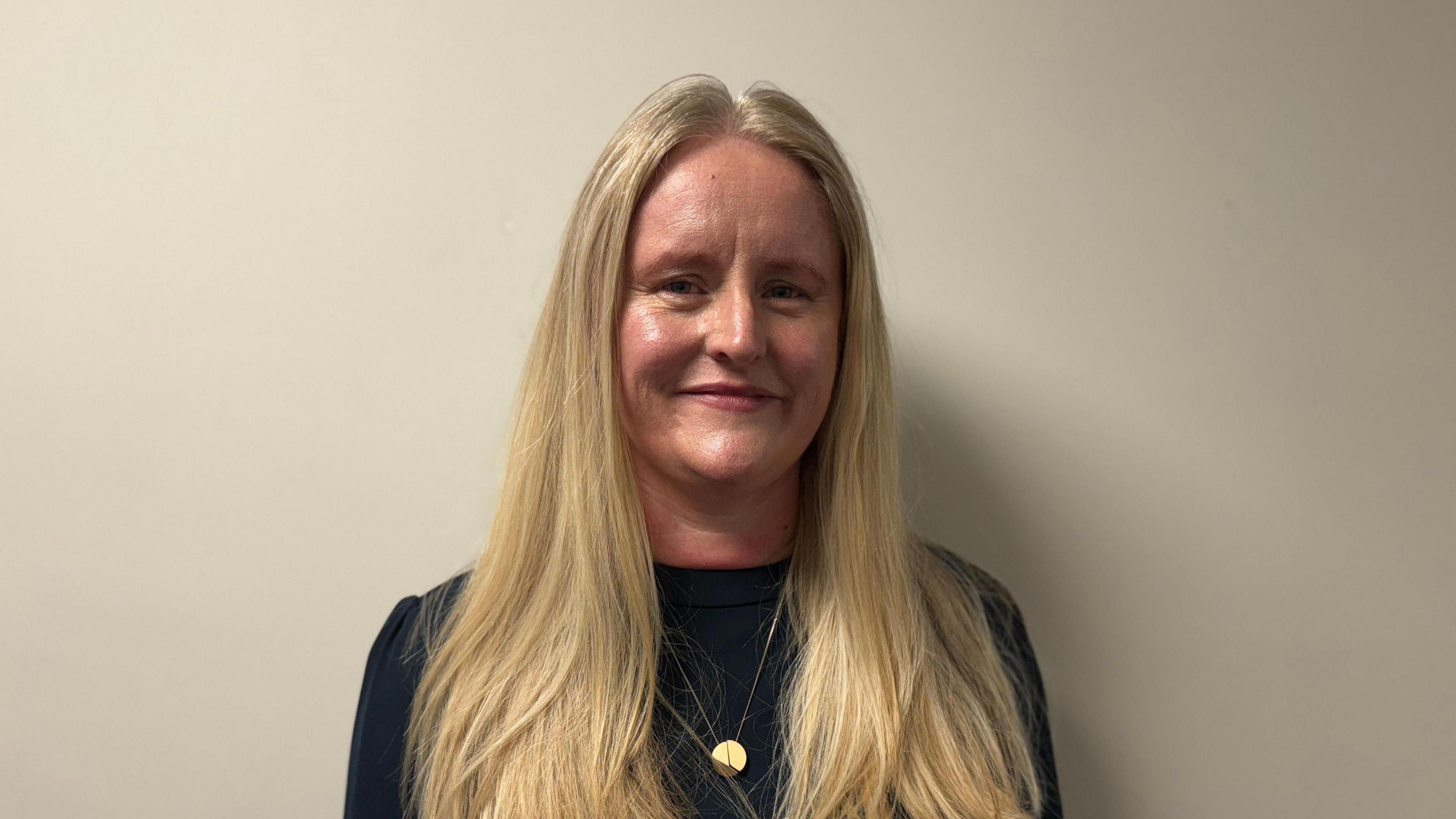 Ms Hanna with long blonde hair wearing a black top as she stands against a white wall.