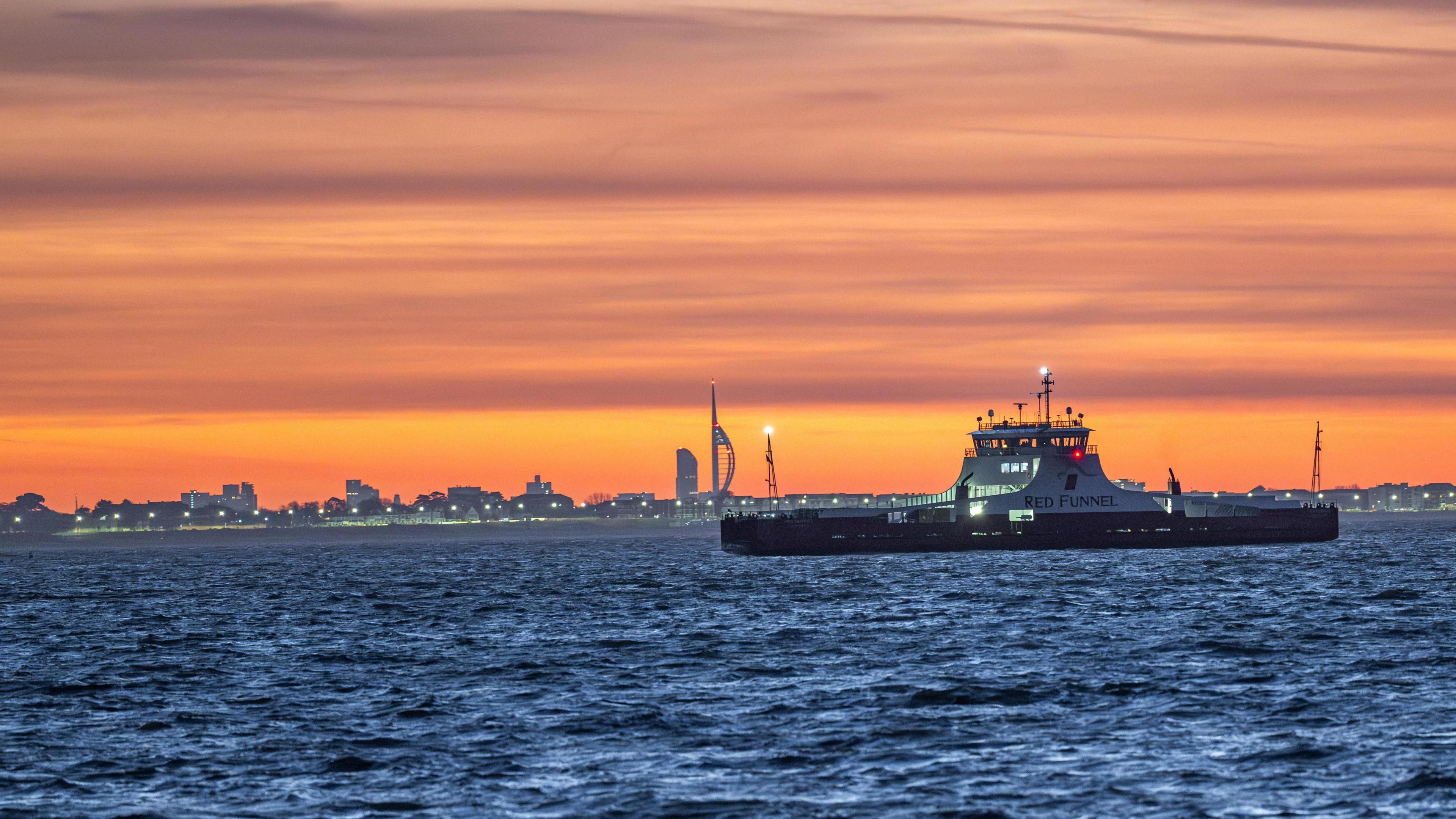 A ferry sails through the shot on the dark blue sea while a bright orange sky fills the rest of the image. An urban horizon is in the distance with the Spinnaker Tower in Portsmouth clearly visible. 