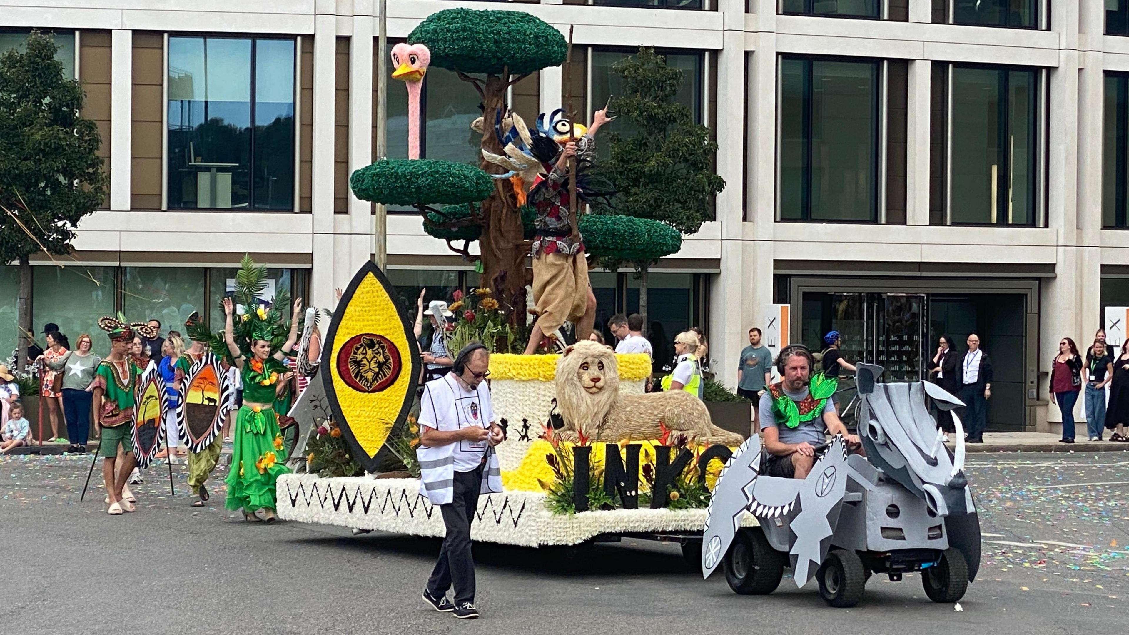 A float featuring a lion, ostrich, colourful bird, African-style shields and a tall tree. It is being towed by what looks like a ride-on mower that is dressed up to look like a rhino. A woman walks behind the float wearing a bright green skirt, top and a headdress which have exotic fruit on them. There are also men dressed in green carrying African-themed shields. There are spectators on the streets.