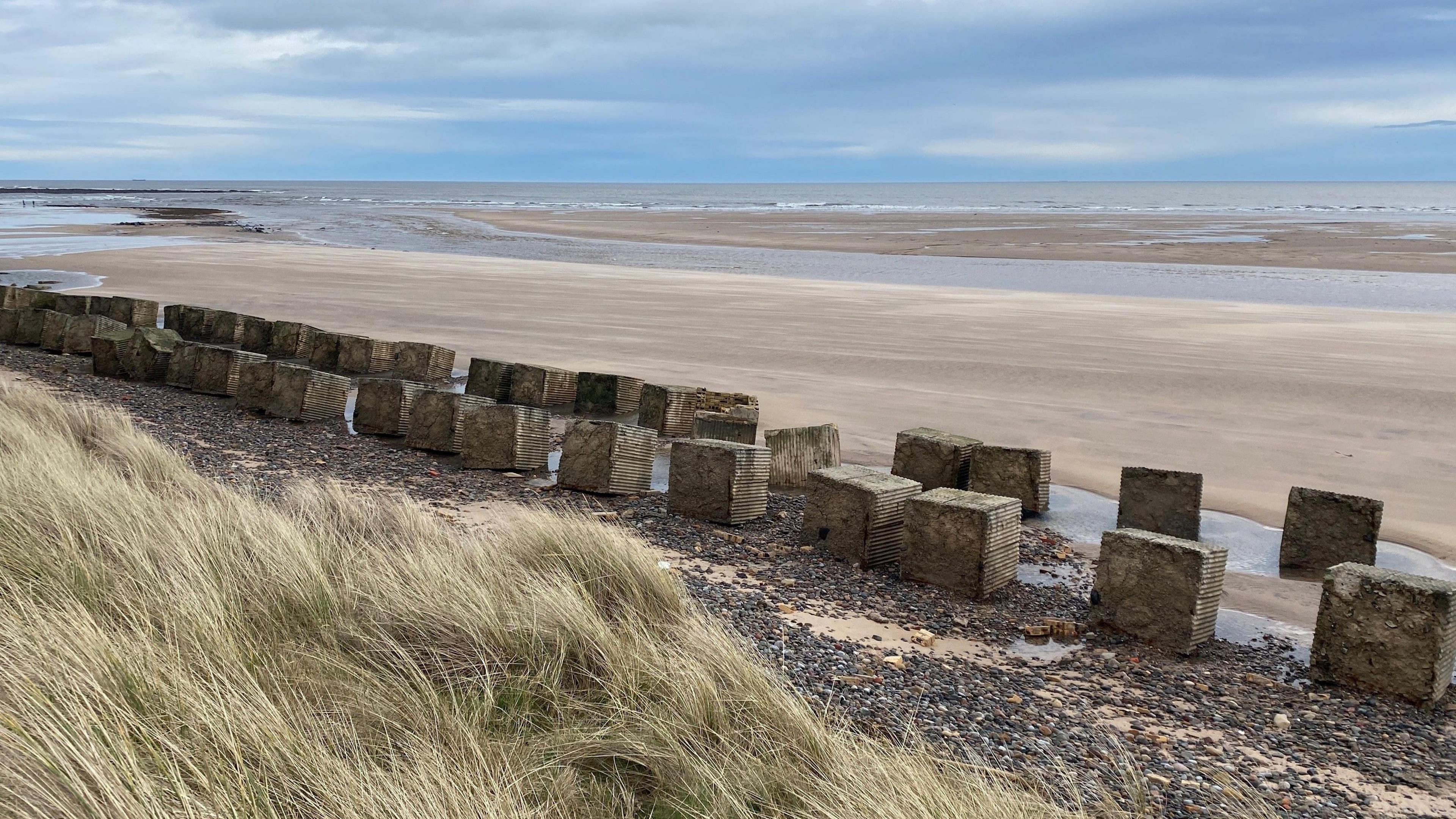 A double row of concrete blocks on a big sandy beach with grassy dunes behind.