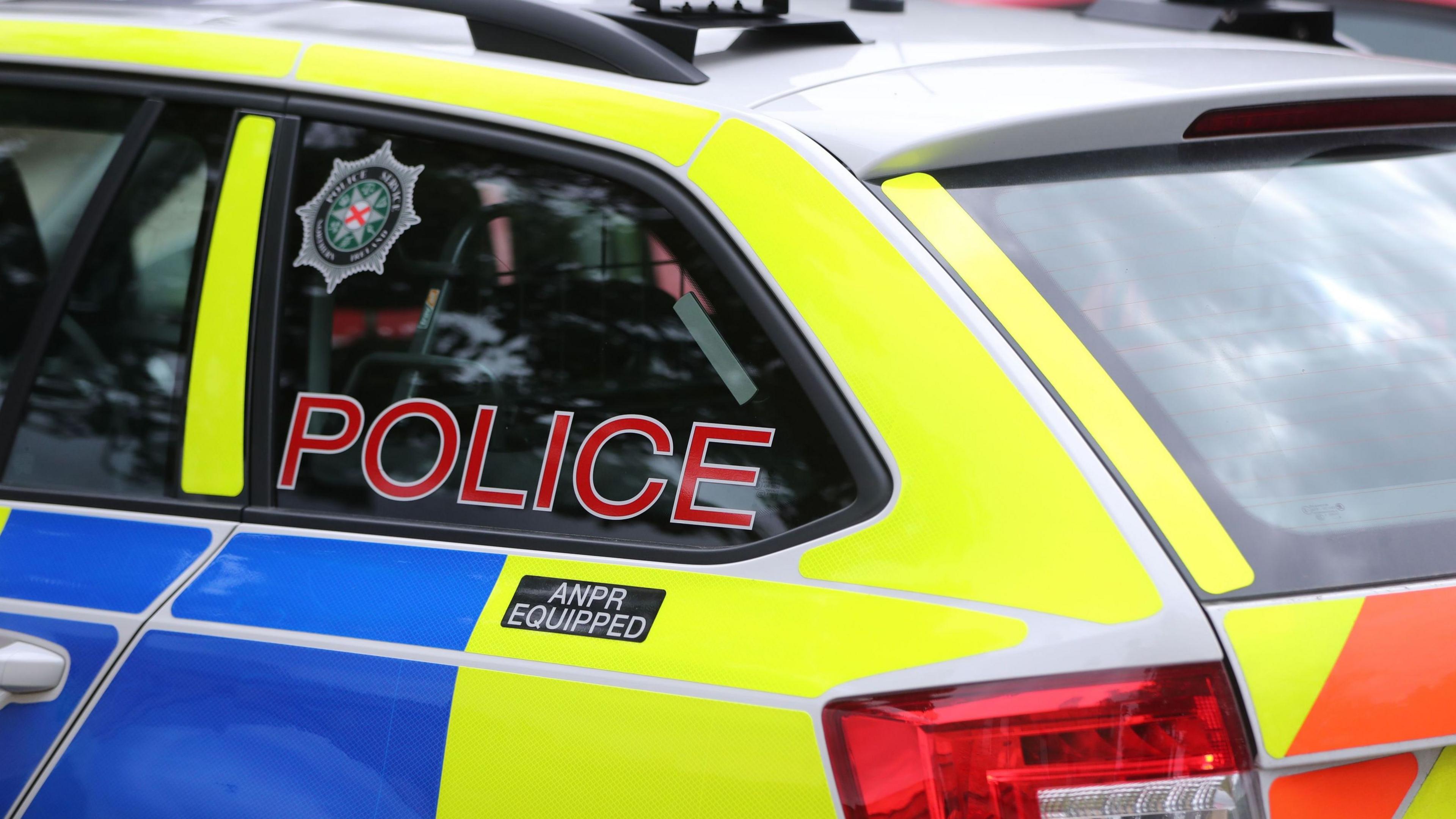 Side view of a PSNI vehicle, coloured in blue, light-green and orange. The window reads police in cpital red letters.