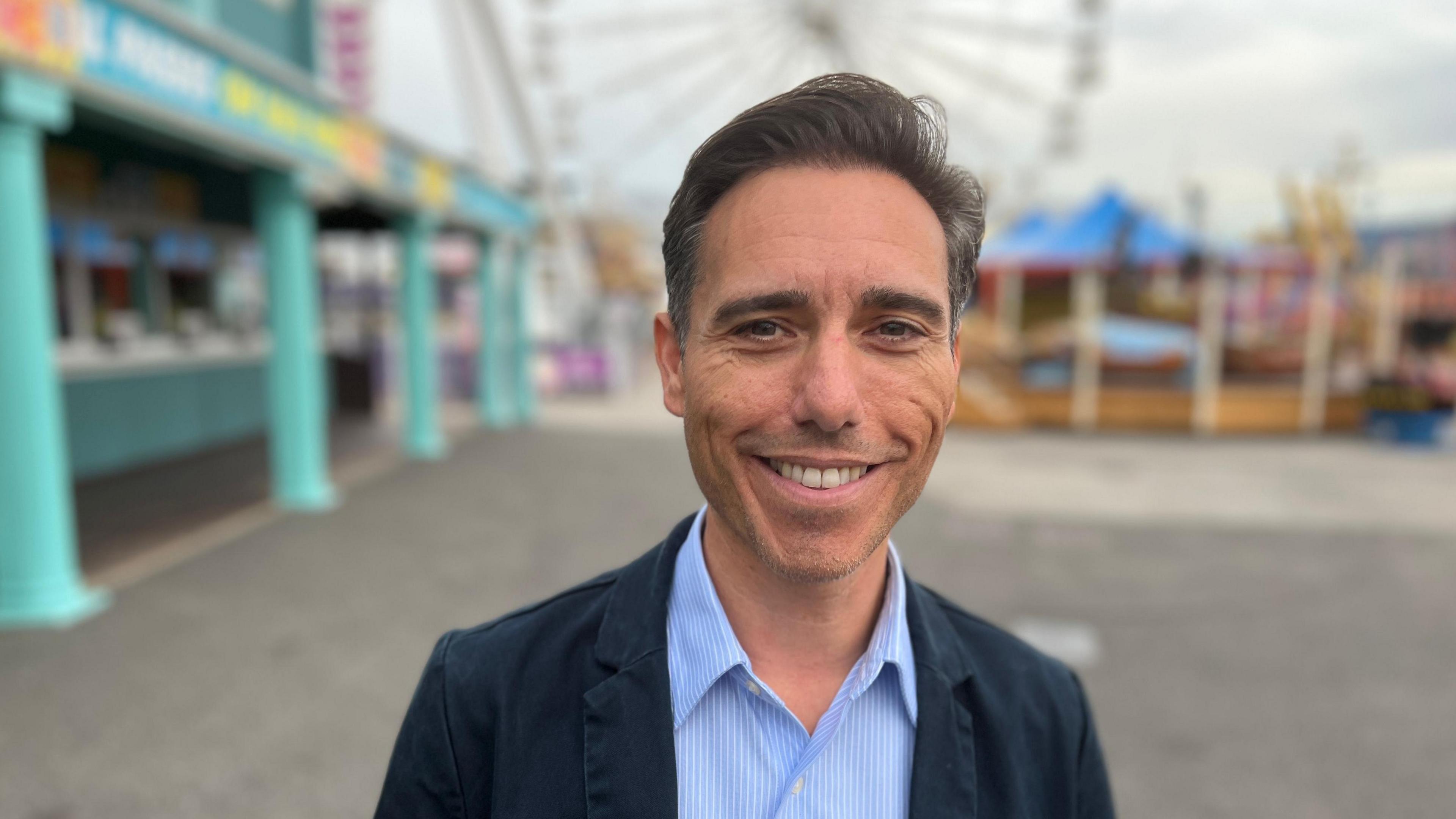 James Miller stands within Adventure Island amusement park in Southend. In the blurred background amusement rides and part of a ferris wheel can be seen. Mr Miller has short dark hair and smiles at the camera. He is wearing a navy suit jacket with a light blue shirt underneath. 