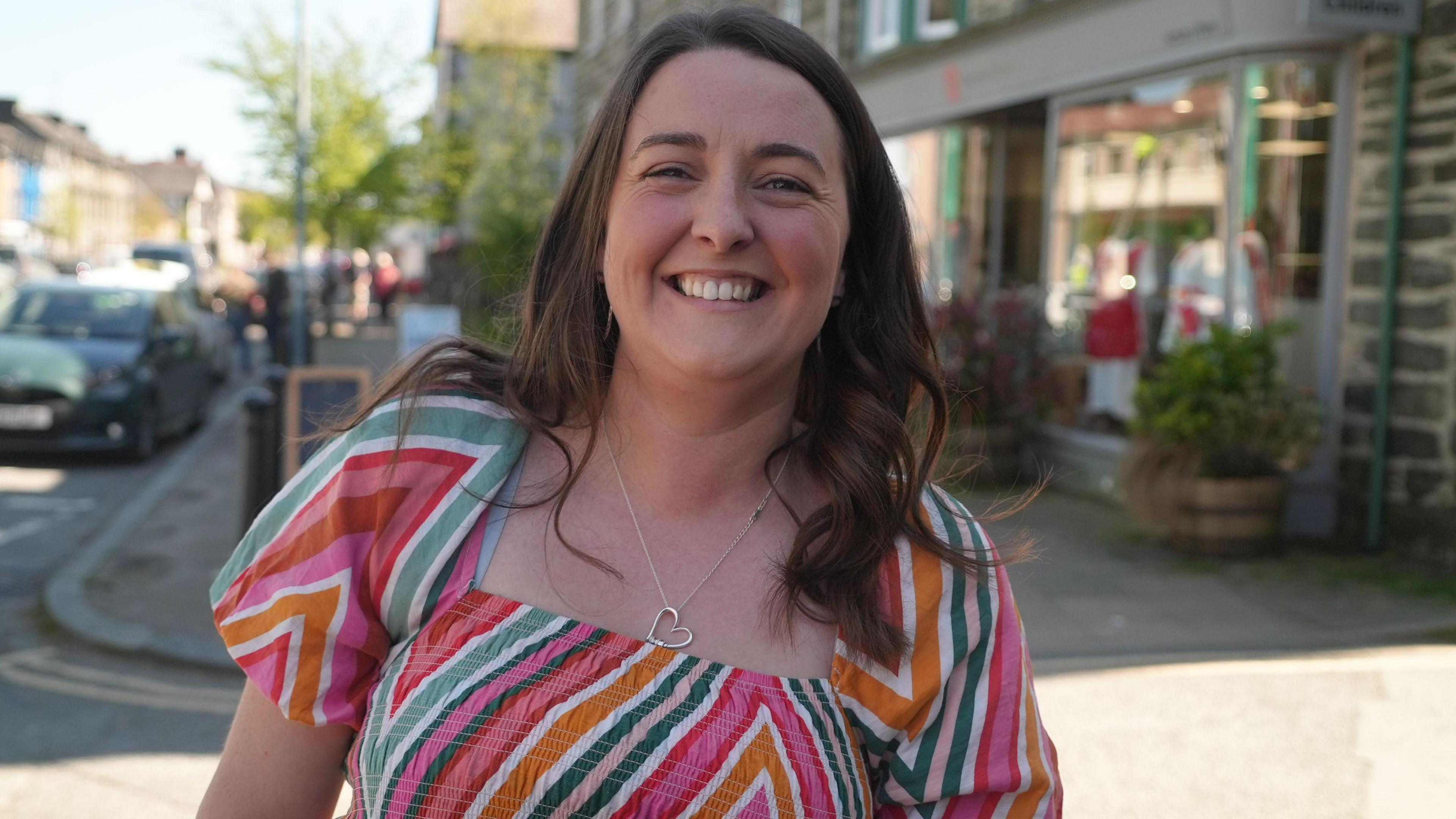 Llun o ddynes ifanc ar y stryd fawr mewn ffrog liwgar. Mae hi'n gwenu ar y camera.