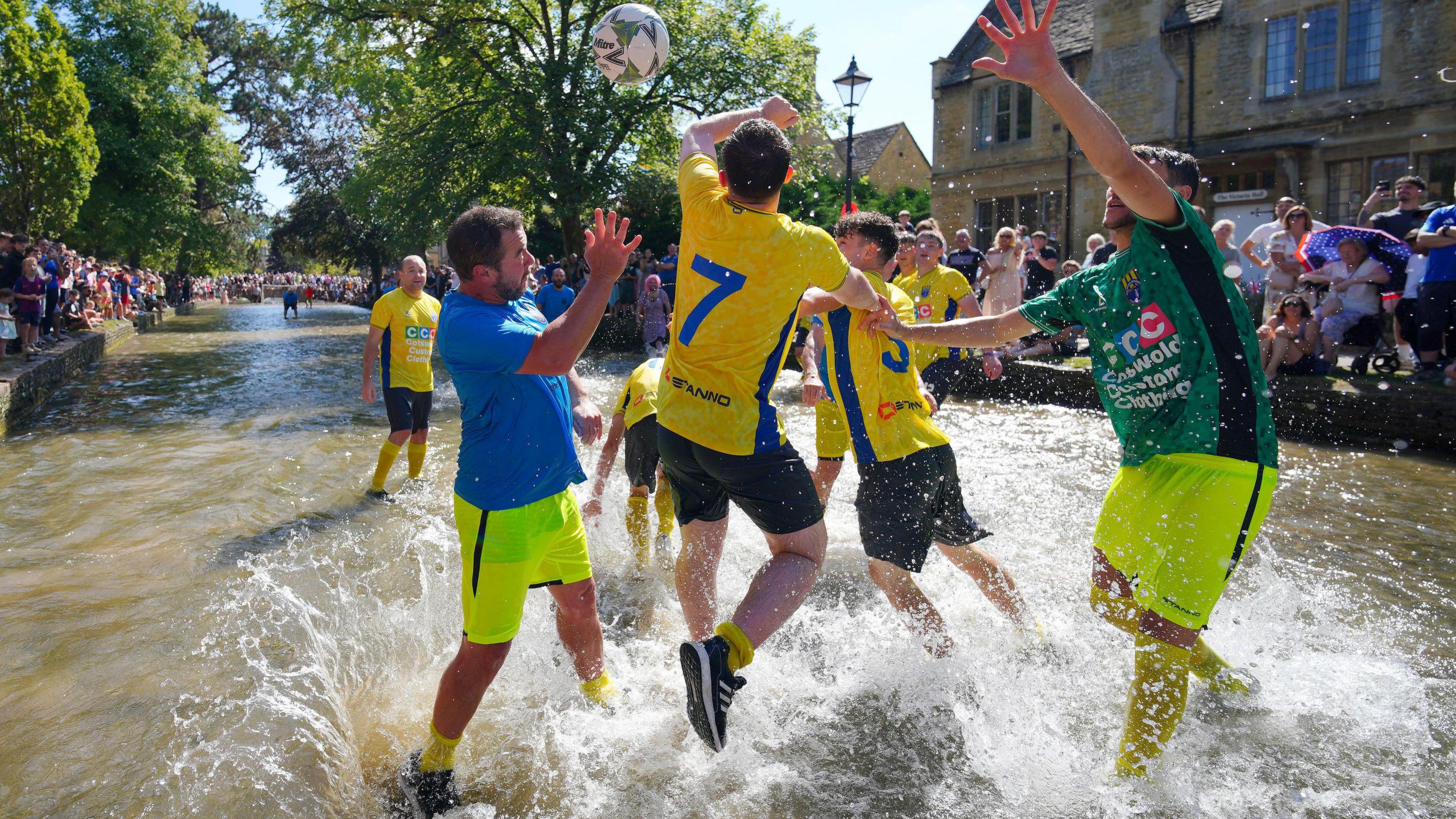 Men playing football in a shallow river, splashing water as they leap for the ball. One team wears yellow and black, the other blue and green. Spectators watch from the riverbanks, with historic stone buildings and a clear blue sky in the background.
