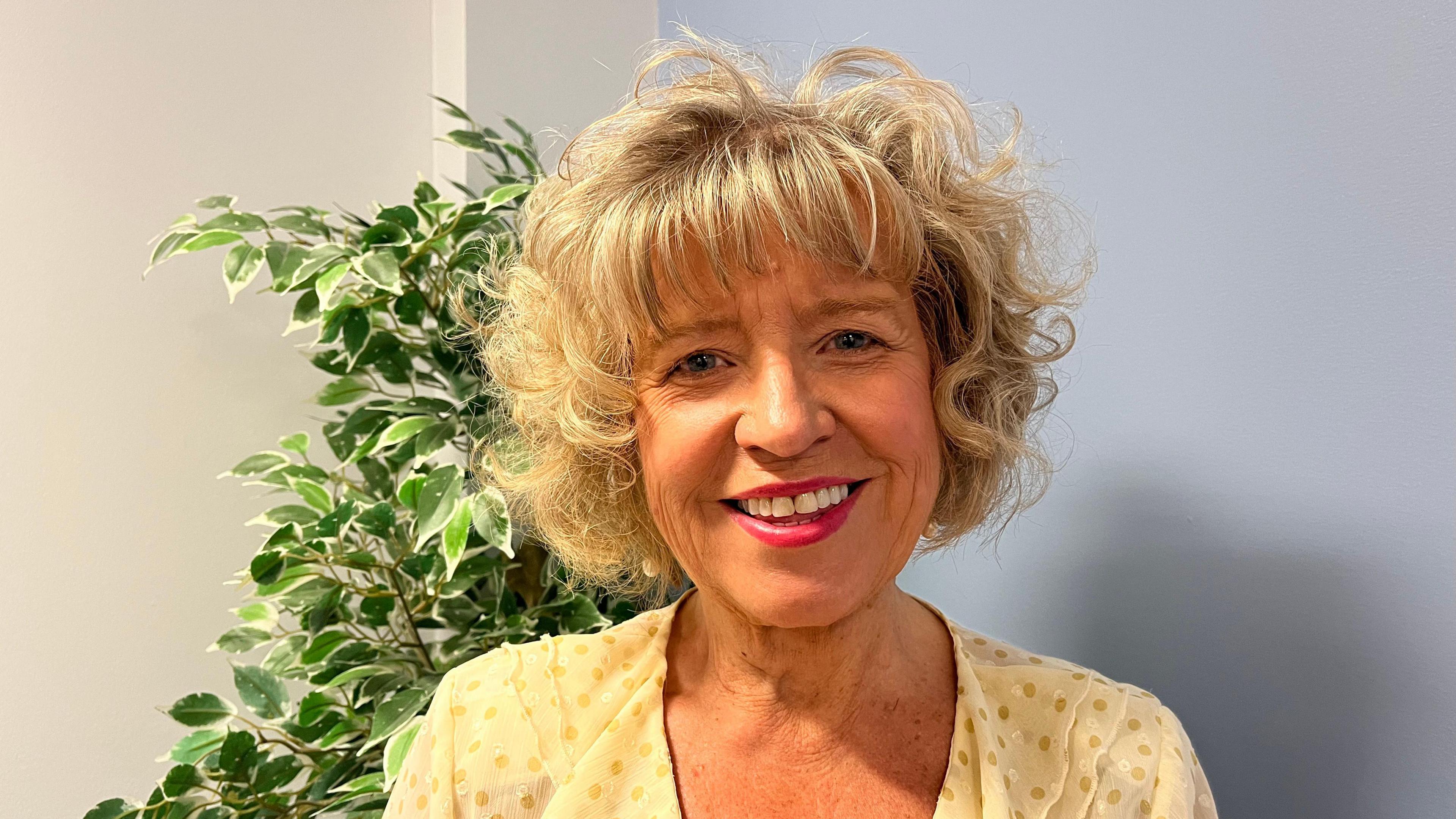 Julia is pictured smiling and looking into the camera. She has short curly blonde hair with a fringe. She's wearing a yellow blouse, and is stood in front of a small tree in a plant pot with green leaves. She's stood indoors, in front of a lilac wall.