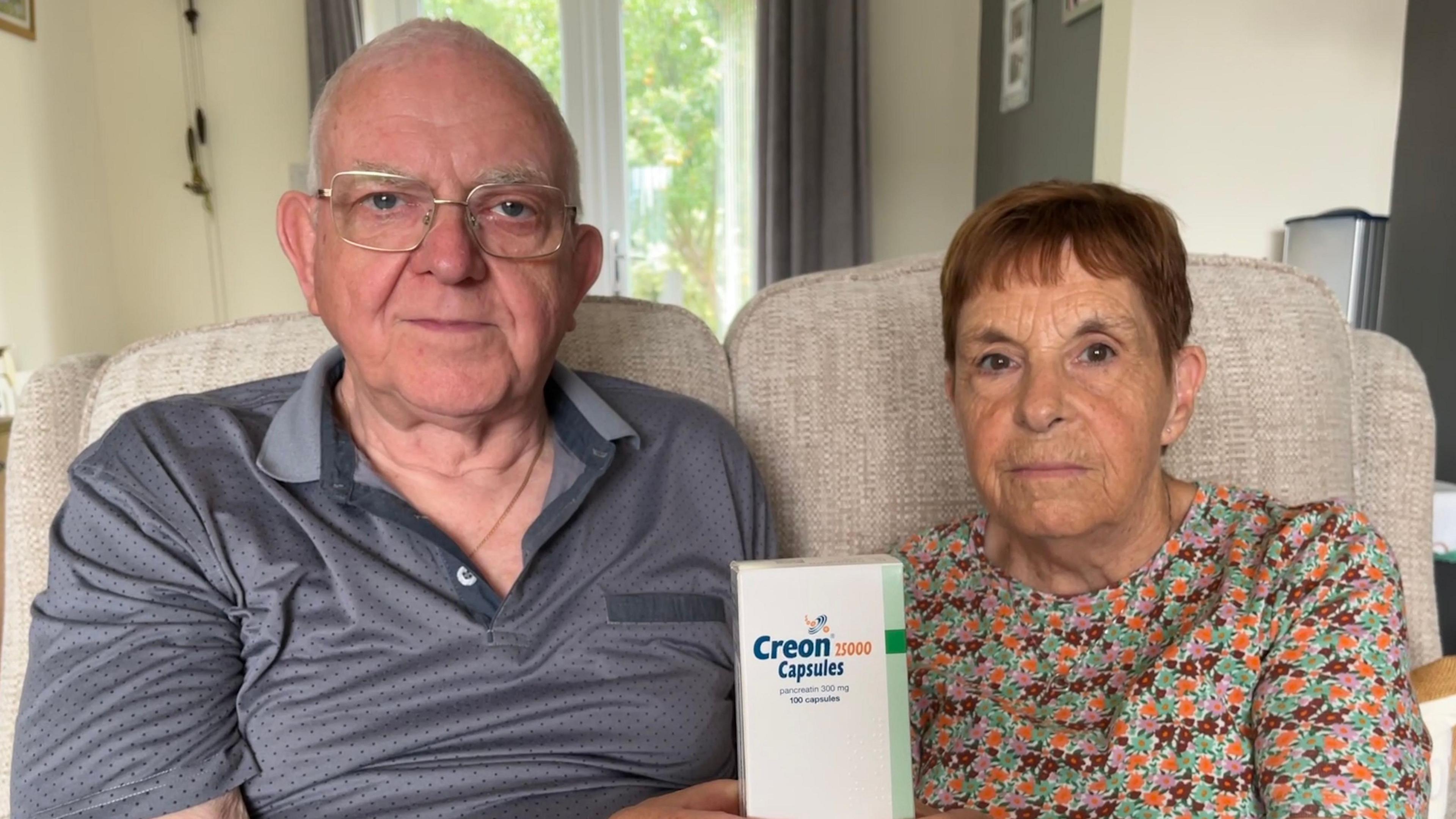 Mr and Mrs Allen sitting on a beige sofa, holding up a box of Creon capsules