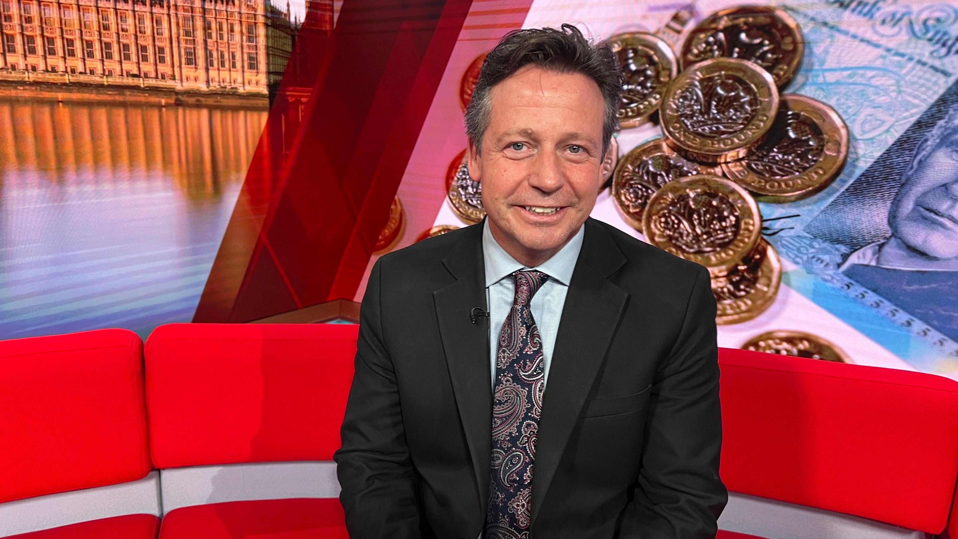 A man with dark brown and grey hair, wearing a light blue shirt, with a black and brown patterned tie and a black suit jacket. He is sitting on a red sofa, with a photo of bank notes and coins behind him