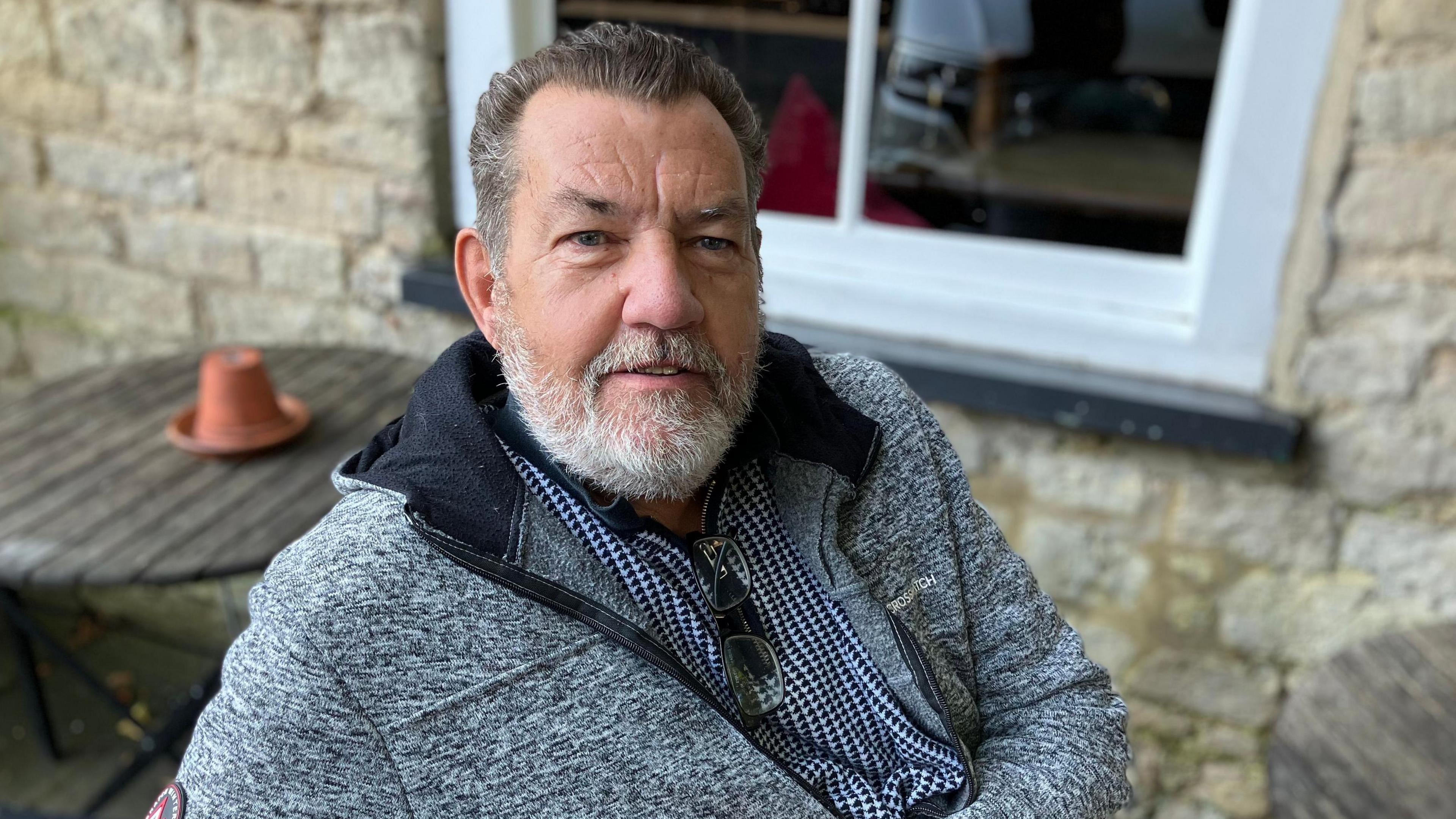 Mark Harris has short brown hair and a greying beard. He is wearing a chequered shirt and a grey top. He is sitting close to a stone building with white framed windows