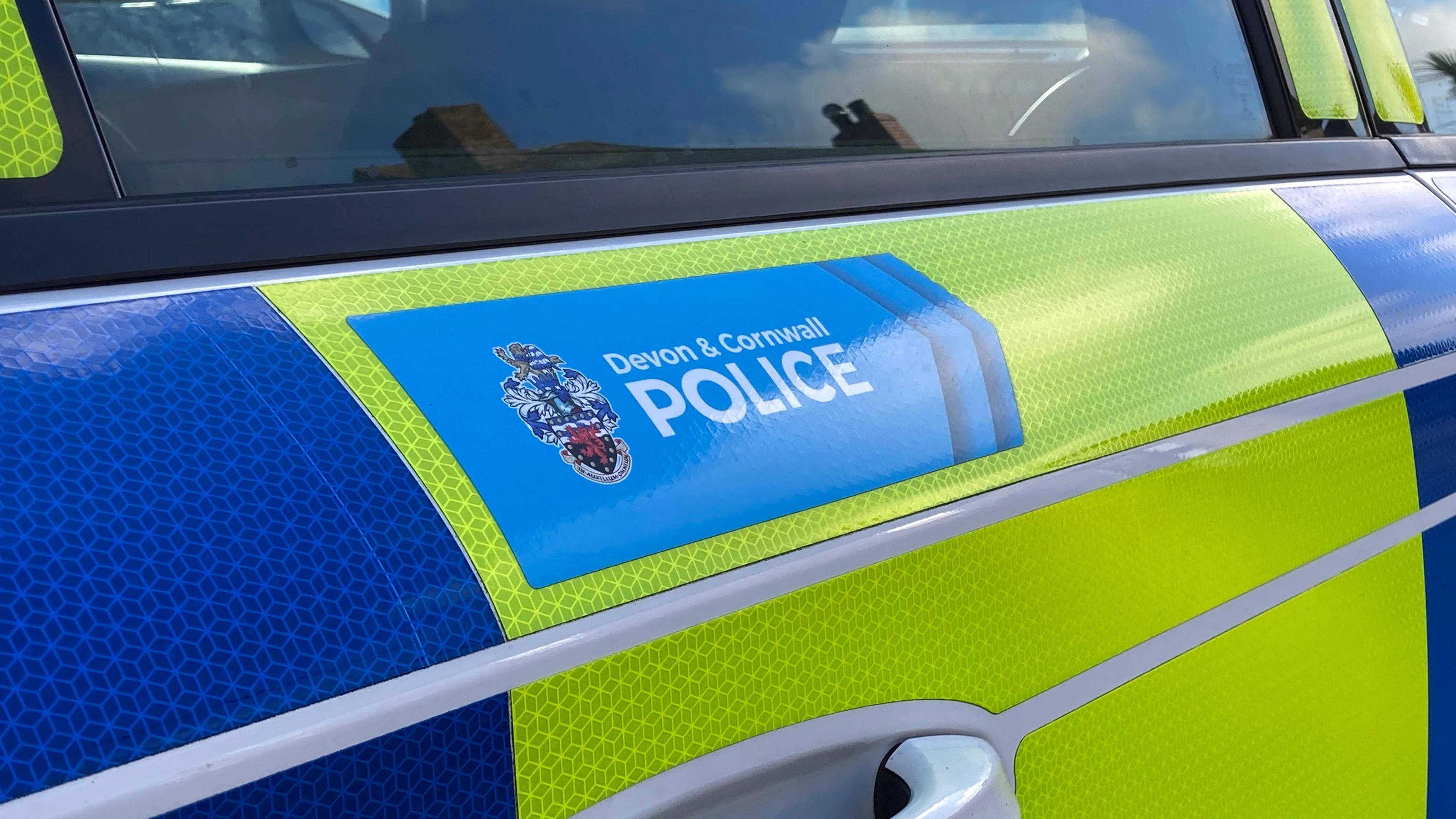 The side of a blue and yellow police vehicle. It has a blue box with a point at the top of the door, below the window, with the words 'Devon & Cornwall Police'. In the window is the reflection of the top of a house.