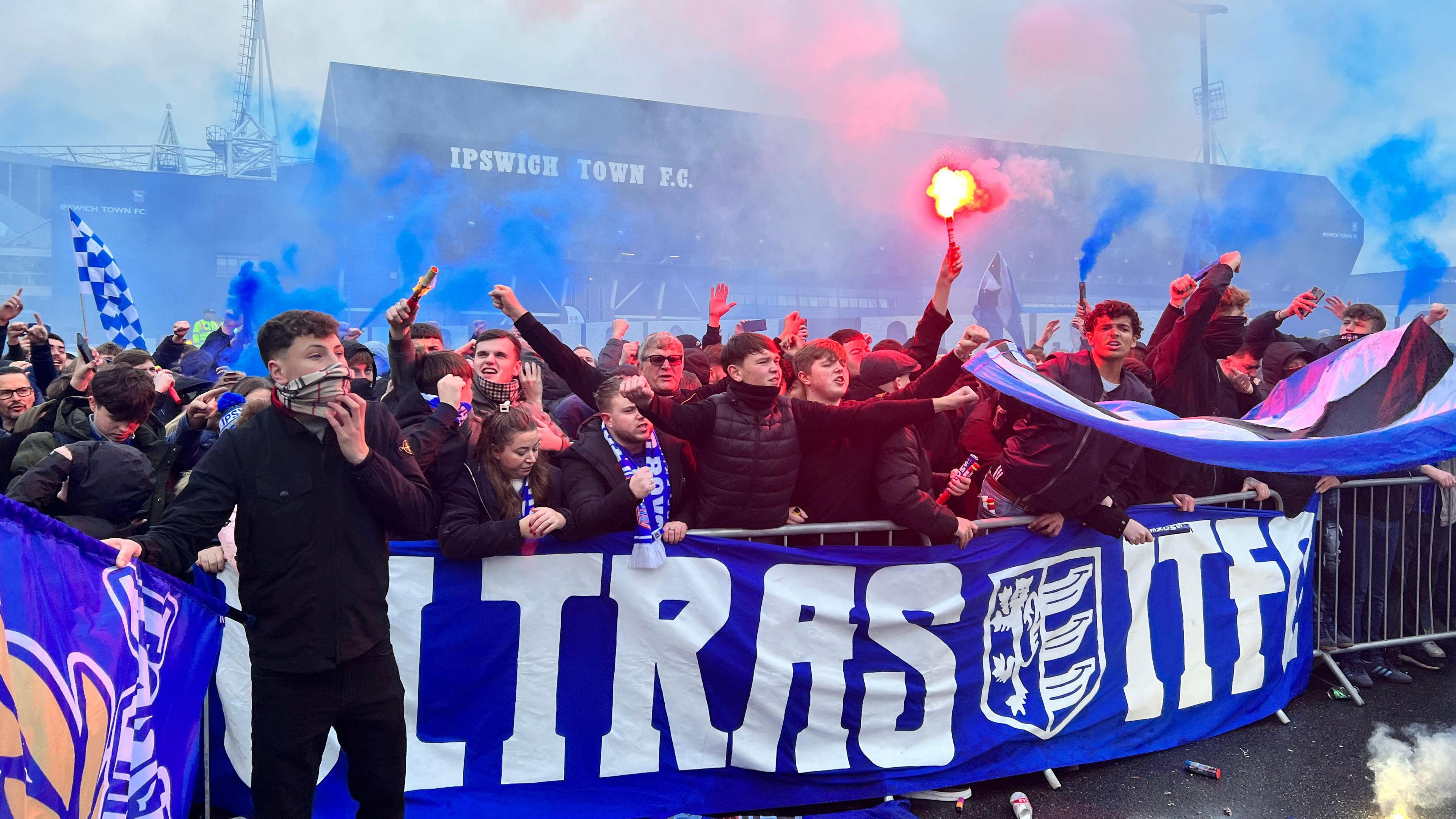 Ipswich Town fans stand behind a metal fence near Portman Road stadium. Some of the crowd wave red and blue flares as well as Ipswich Town flags.