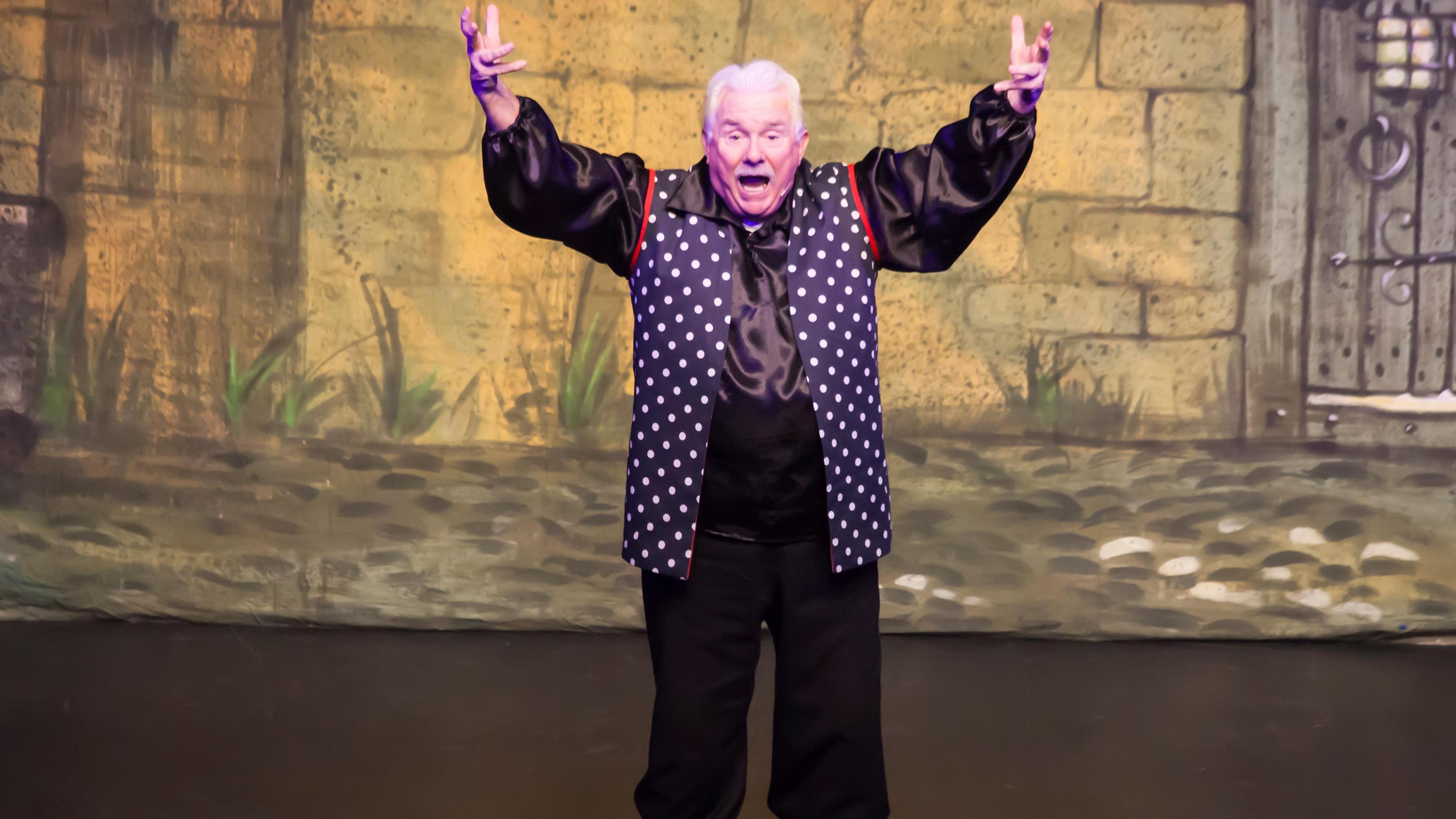 Owen Money on stage with arms up in the air and mouth open in wide black trousers and navy waistcoat with white polka dots and a red trim over a satin black shirt