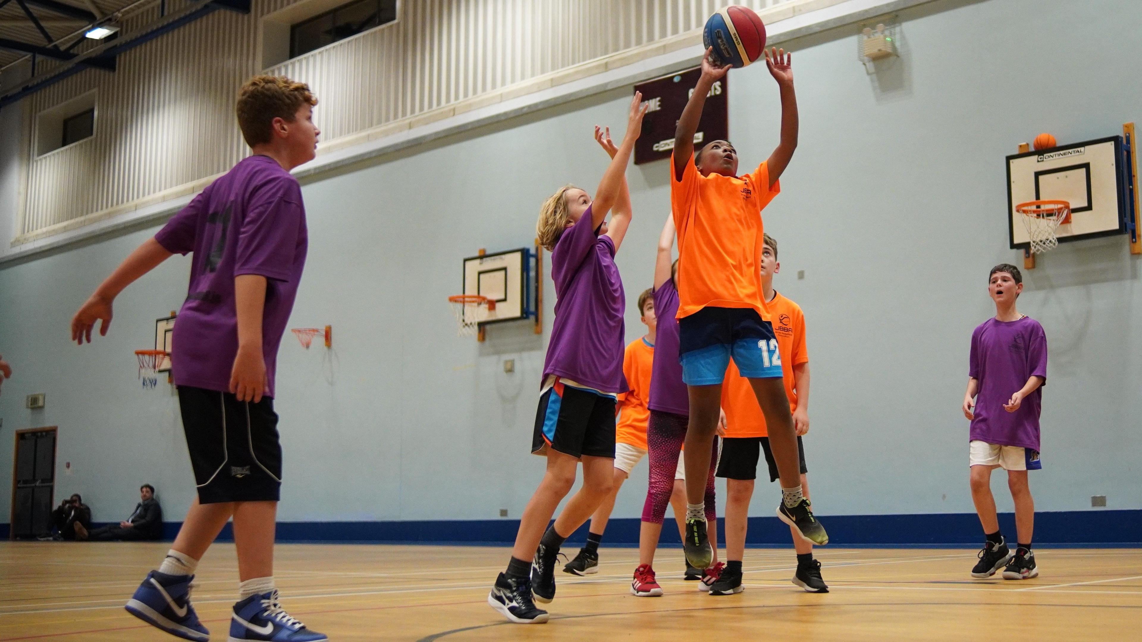 A group of seven children on a basketball court running towards and jumping up to grab a basketball.