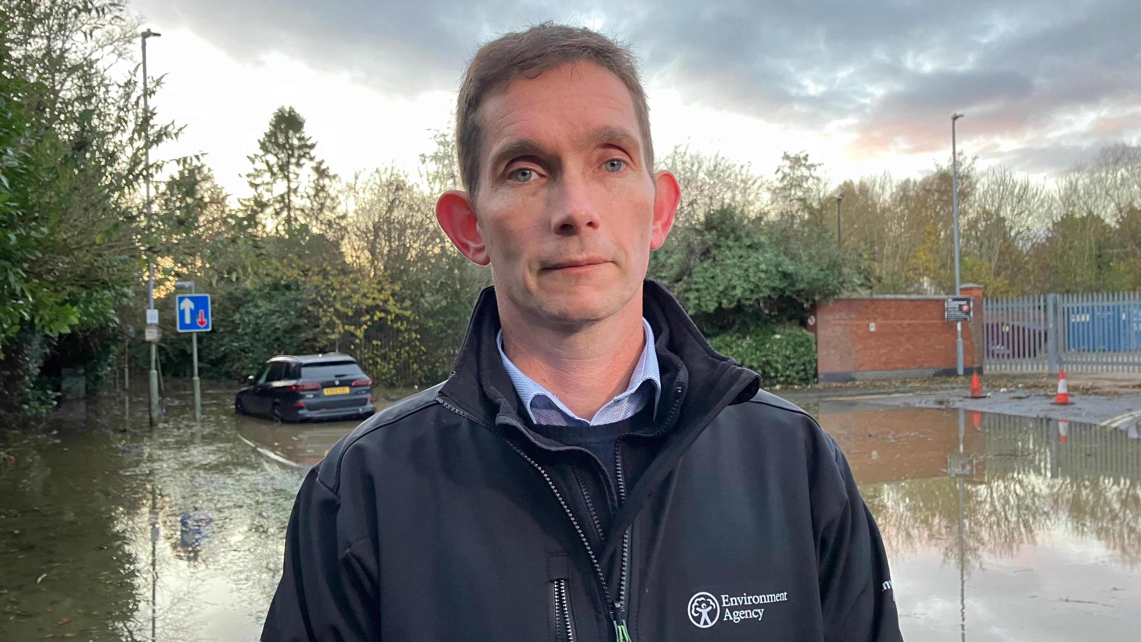 Ben Thornley, from the Environment Agency is wearing a blue jacket, shirt and pullover. He is standing in front of a flooded road and behind him, a car can be seen partially submerged up to its bumper