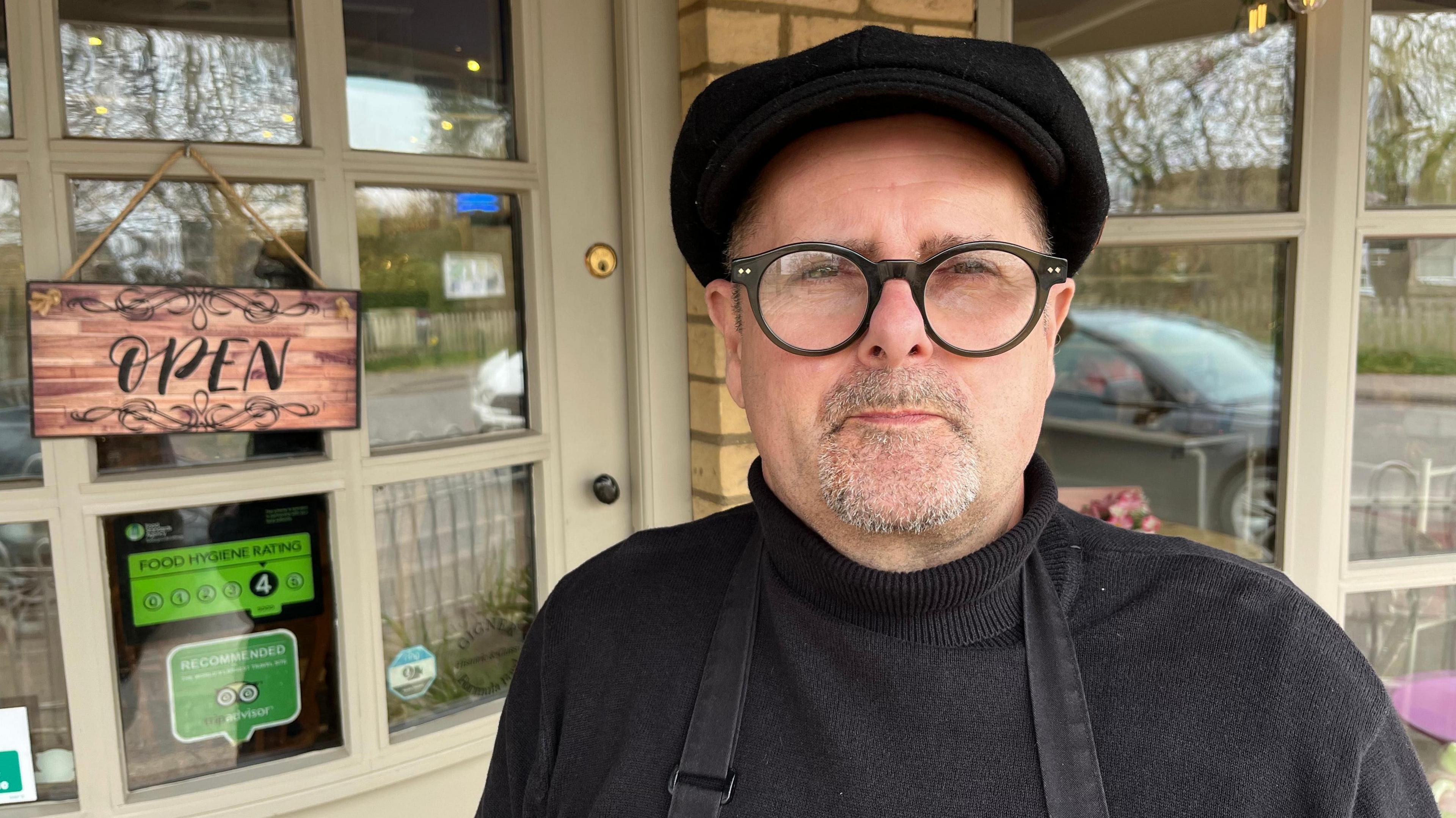 Steve Gigner, 59, is wearing a black flat cap and black jumper, with a black apron over the top. He has a greyish stubbly beard and moustache and is standing in front of the shop door, which has an open sign on it.