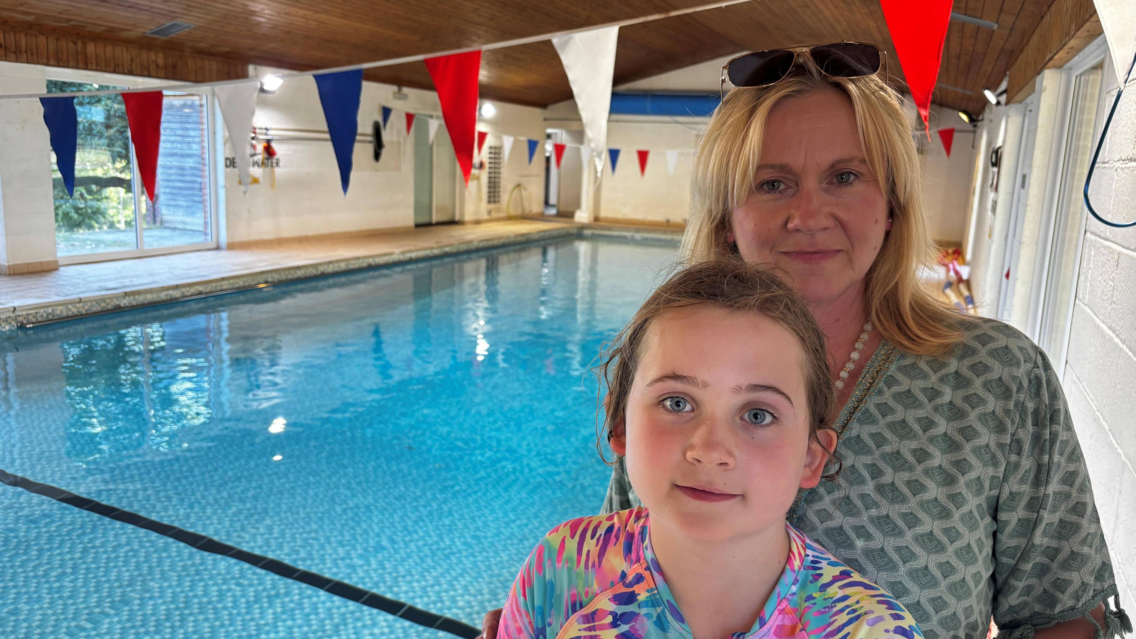 Image shows a pool setting with bunting attached to the ceiling.
In the foreground of the shot is a young girl wearing a multi coloured swim suit with her hair tied back.
Behind her is a woman wearing a green short sleeve dress. She has shoulder length blonde hair with sunglasses on her head. 