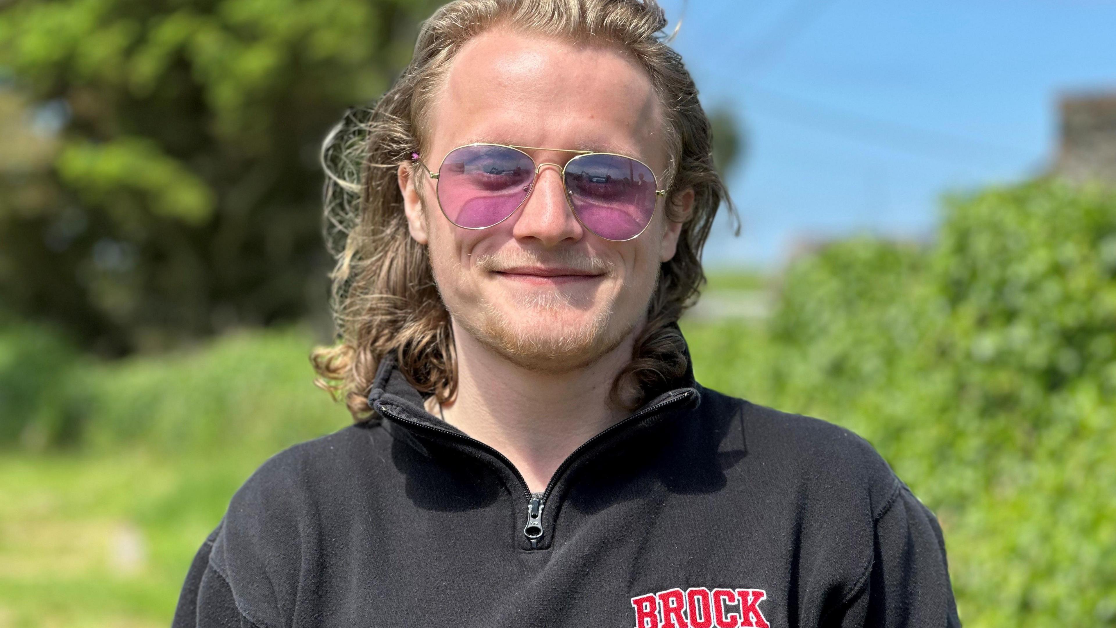 A young man with shoulder length blonde hair. He is wearing a black top with Brock written on it in red and pink tinted sunglasses.