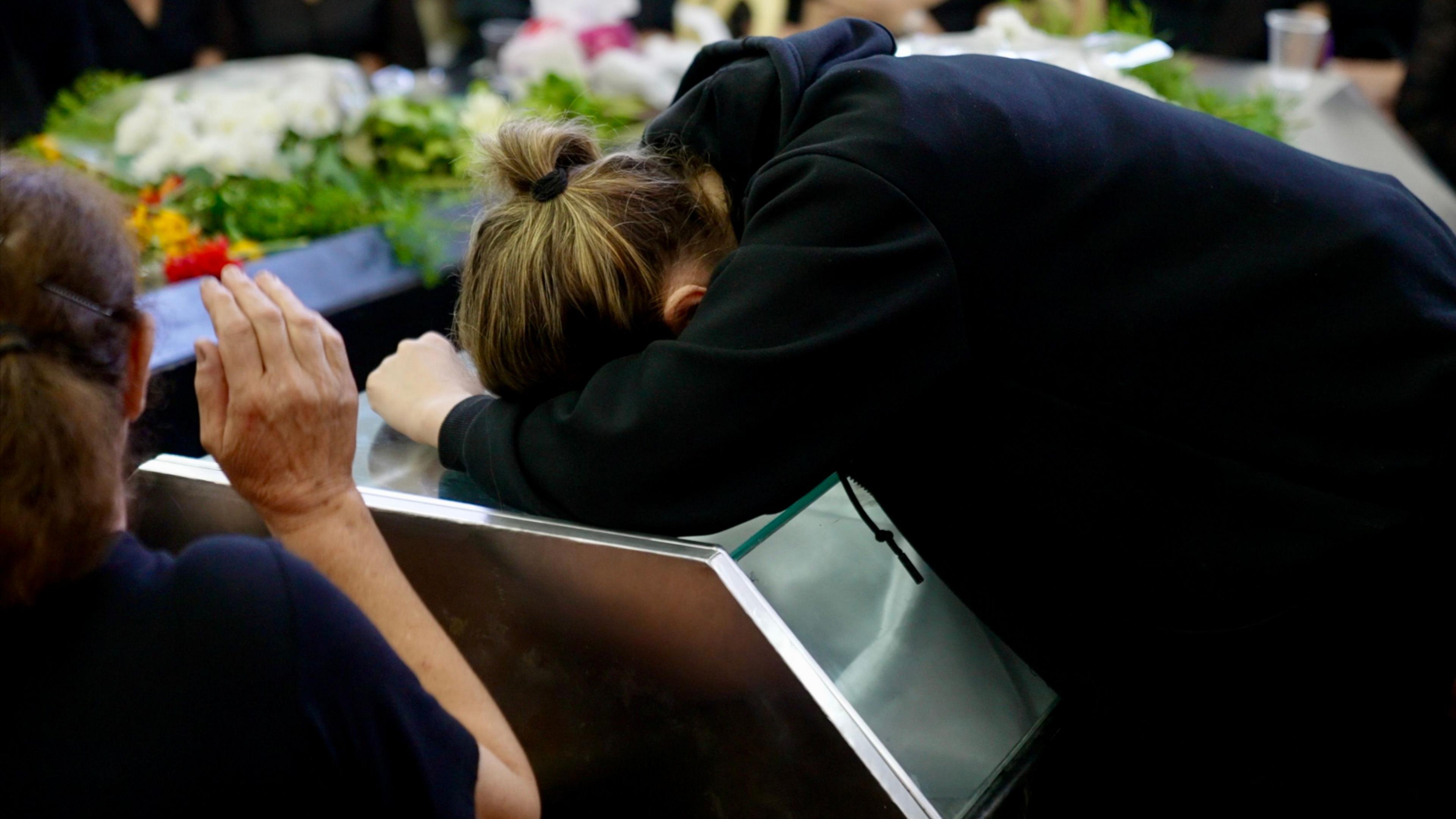 A woman dressed in black with her hair tied back rests over a coffin, head down