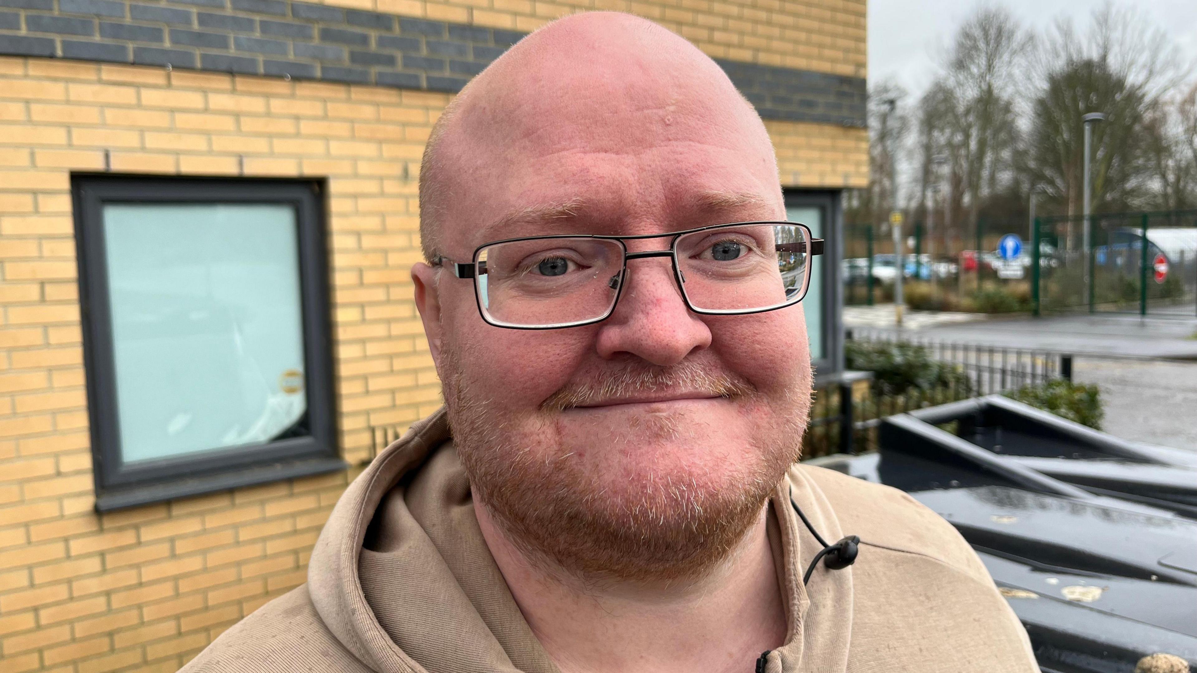 Gareth Lingwood is bald with a thin red beard and moustache. He is wearing glasses and has light brown fleece top on with a hood folded on his neck. Behind him are black bins and a window from the block of flats.