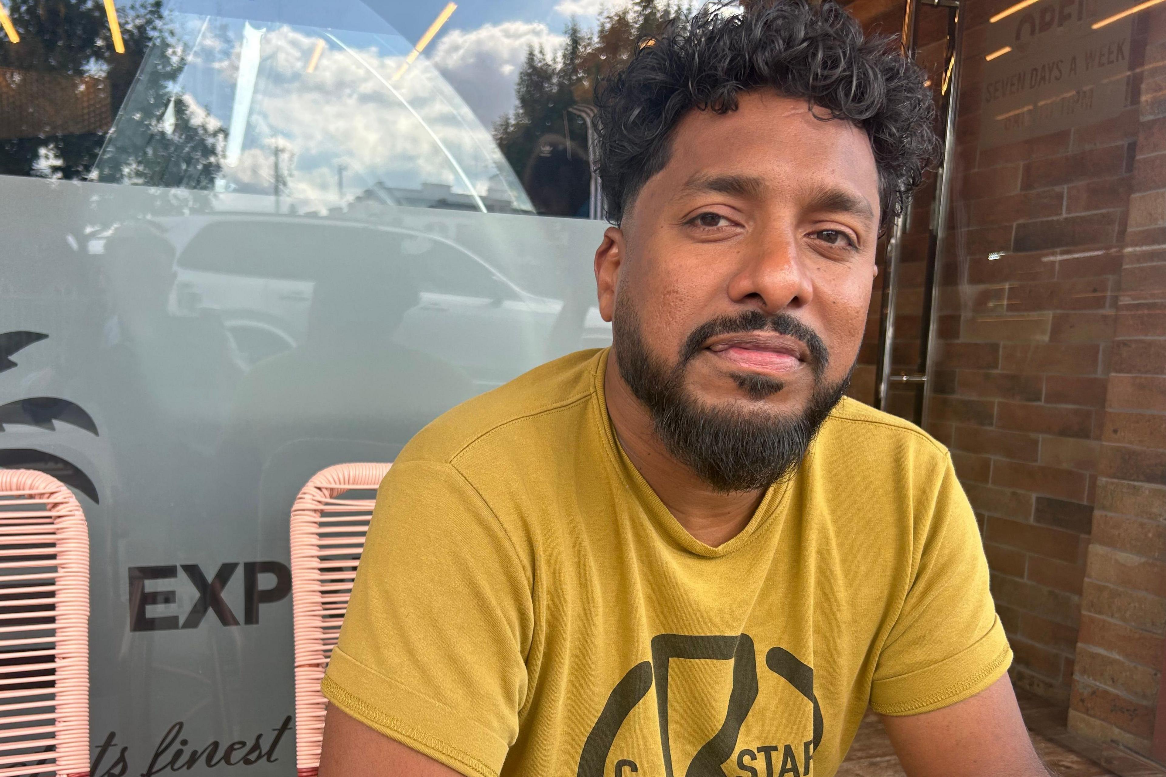Dina Michael who has dark hair and a beard sits on a chair in front of a shop window. He is wearing a yellow T-shirt