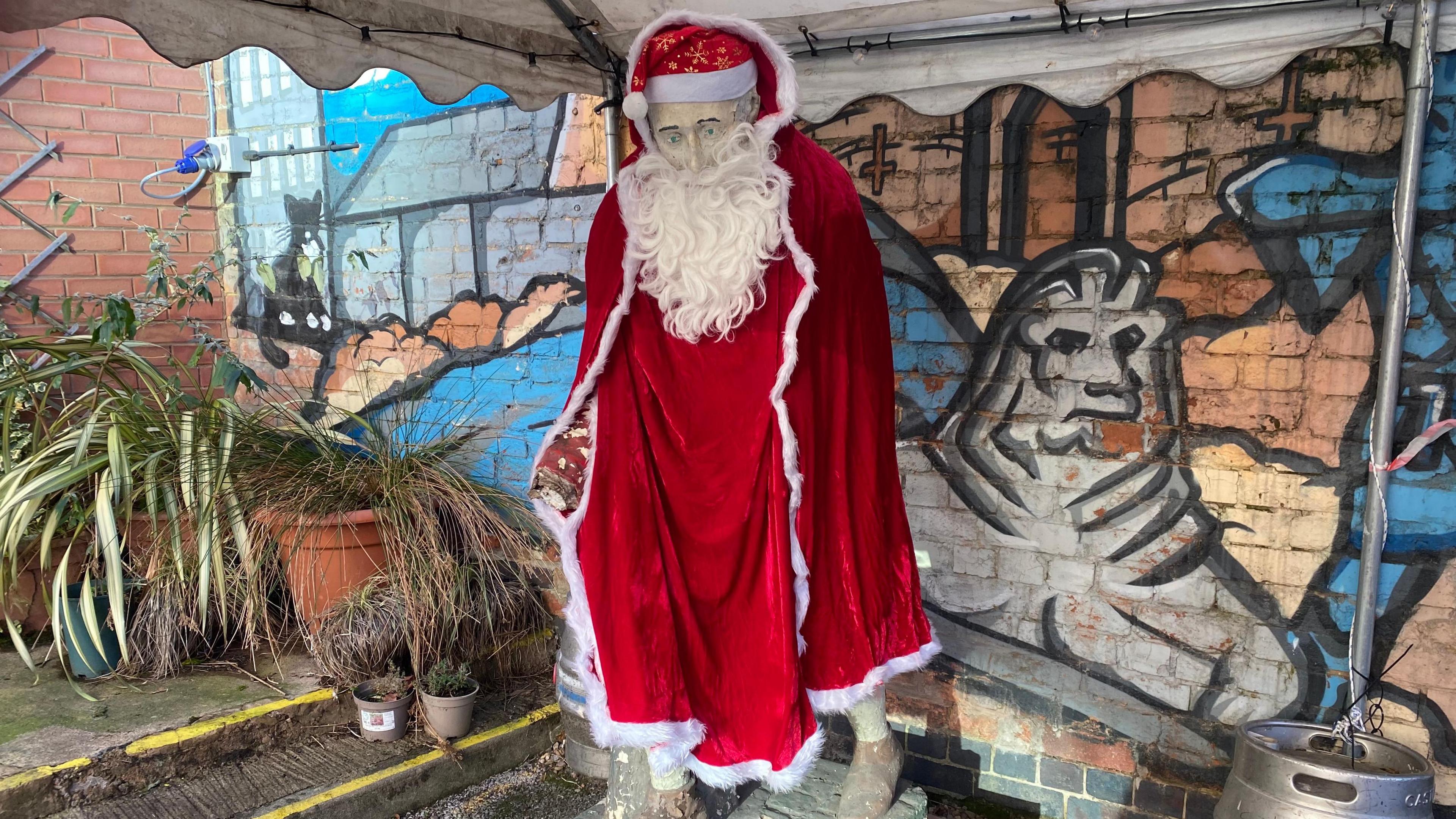 The old general statue wearing a red velvet robe with white fur trim. It has a red Santa hat with gold snowflakes on its head. It is also wearing a long white beard.