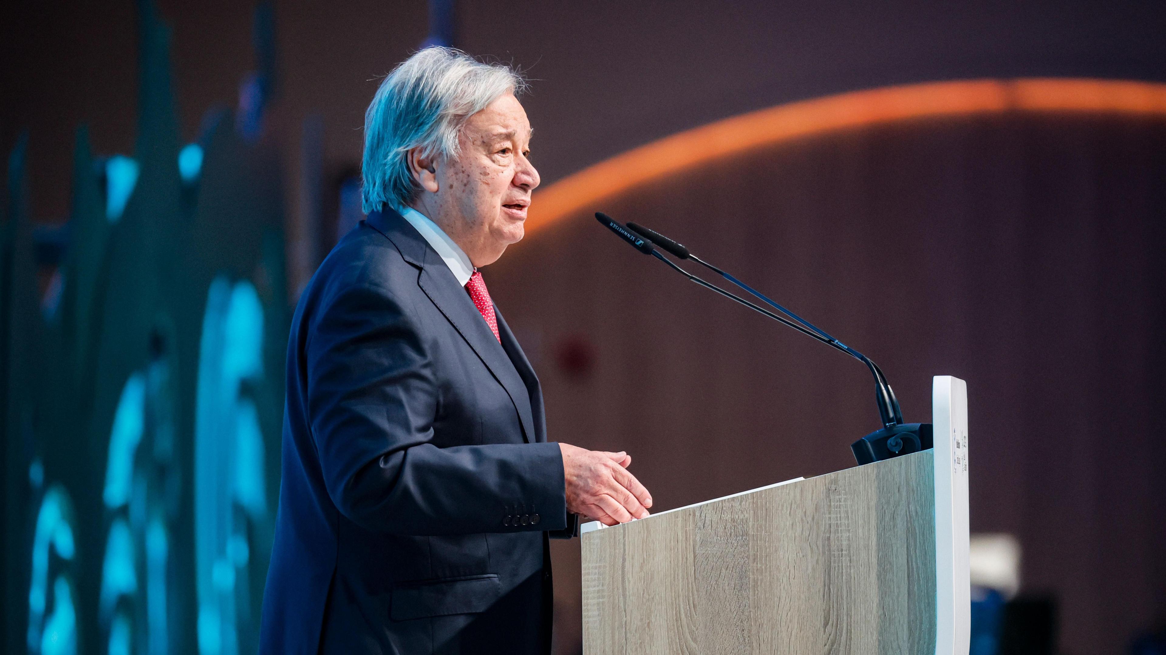 United Nations Secretary-General Antonio Guterres stands at a lectern. He is wearing a dark blue suit and a red tie.