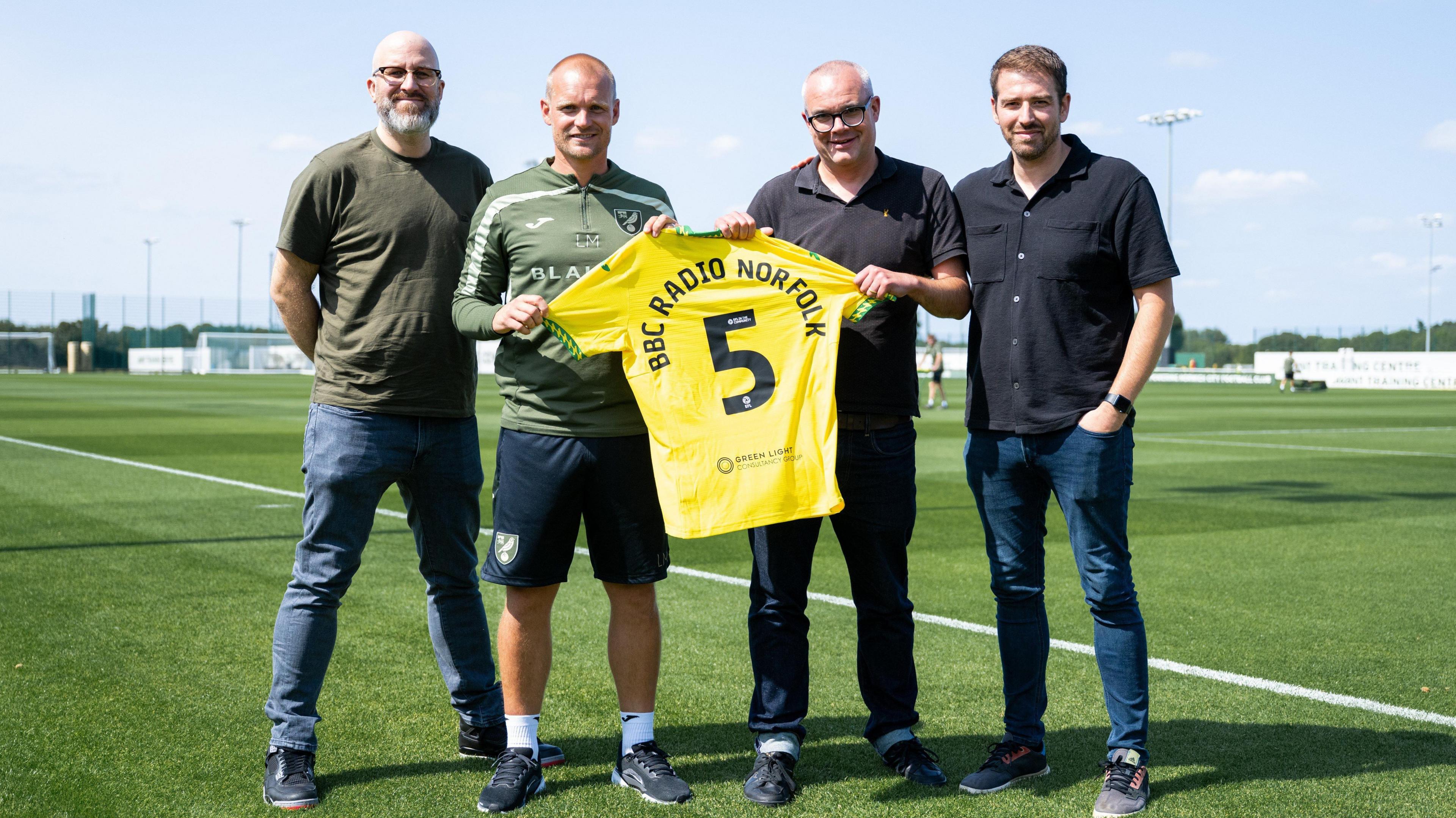 Four men standing in a line and looking at the camera. The two men in the middle are holding a yellow football shirt, with the number "5" and "BBC RADIO NORFOLK" printed on the back. They are standing on a football pitch.