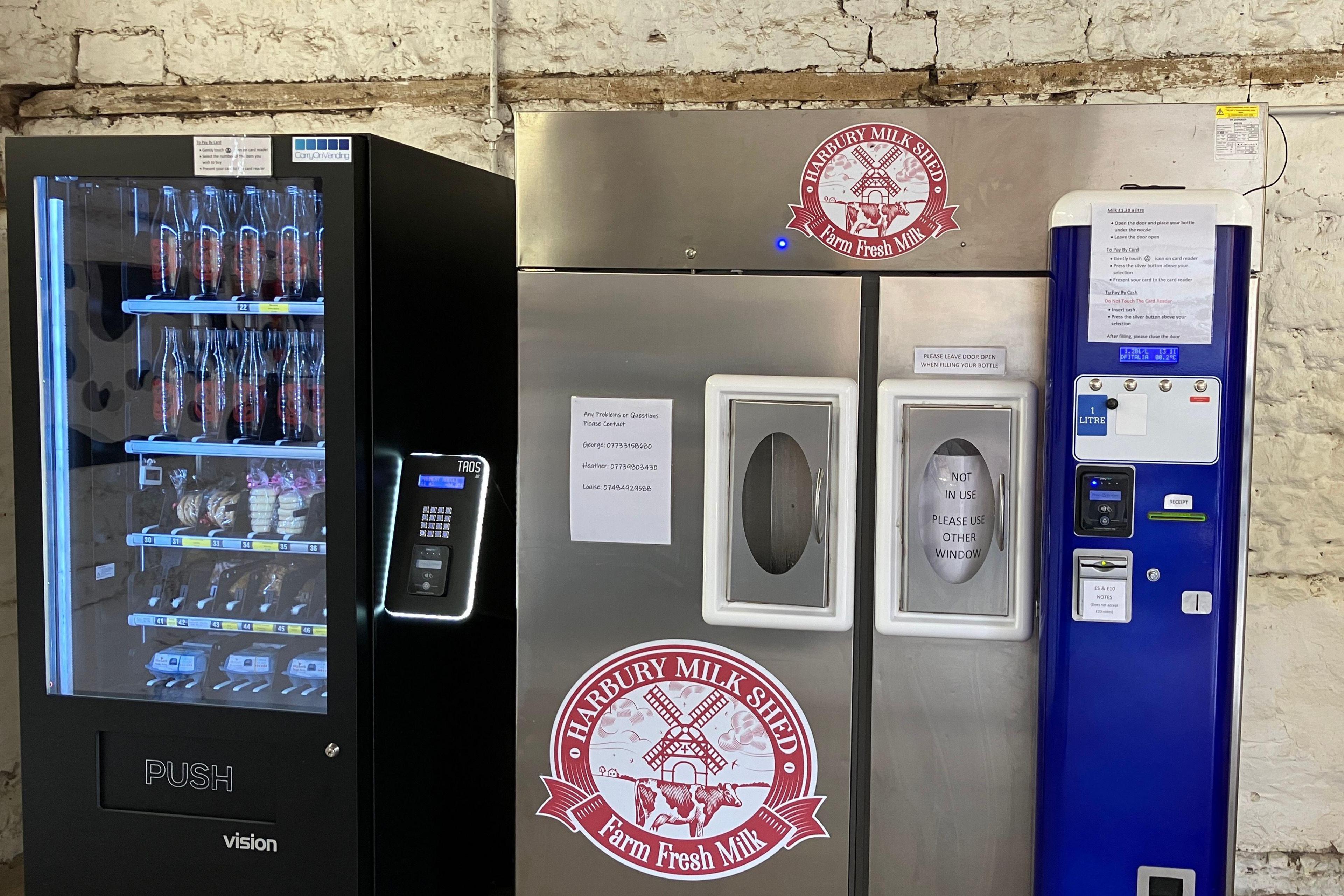 A vending machine is stocked with clean clear glass bottles. Next to it a silver dispenser has a red and white sticker with the Harbury Milk Shed logo. 