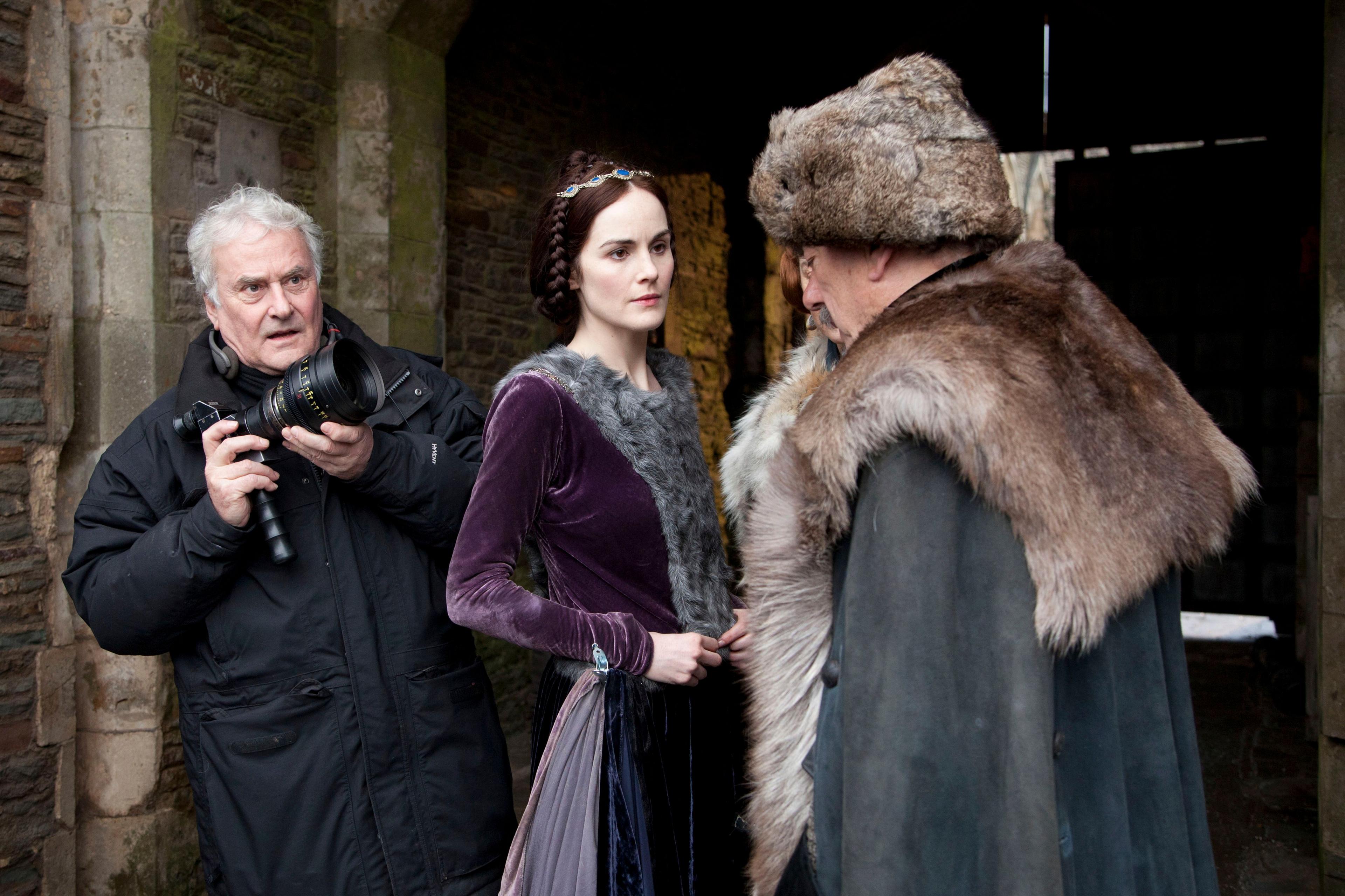(L-R) Richard in dark blue anorak holding a camera, Michelle Dockery in purple velvet dress with grey fur in the centre, looking at Alun Armstrong (side profile) in brown fur hat and far round his shoulders and back and grey cloak