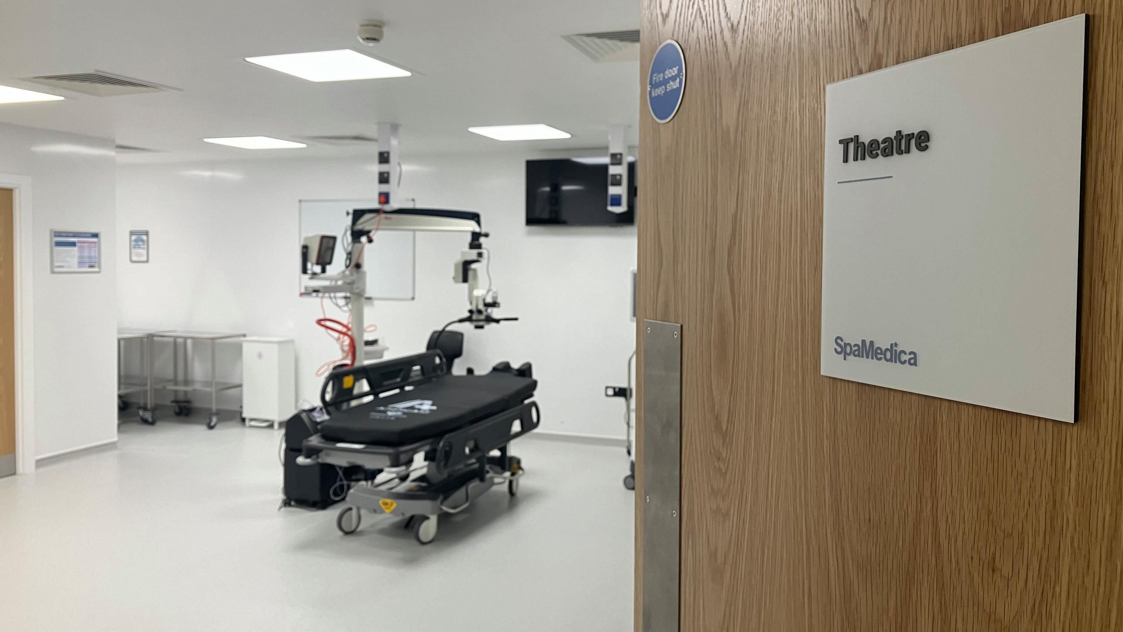 a door to an operating theatre ajar with a sign reading 'theatre spamedica' and a theatre bed and eye surgery equipment