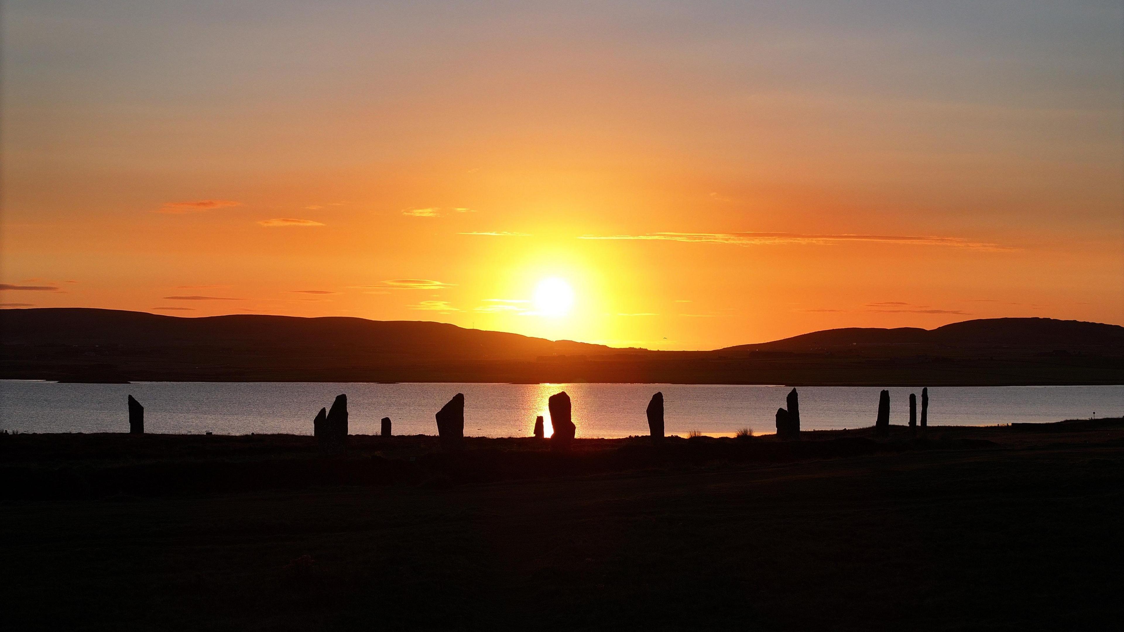 An orange and yellow sunrise behind the silhouette of several standing stones