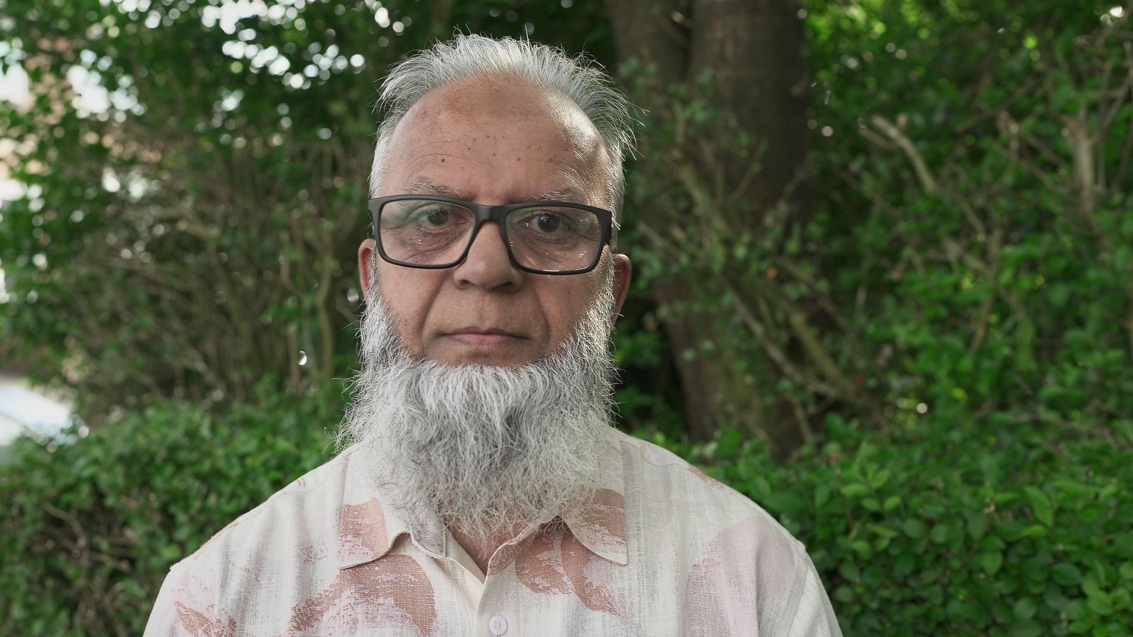 Farooq Khan staring into the camera, wearing a white and pink shirt and glasses, in front of some green bushes