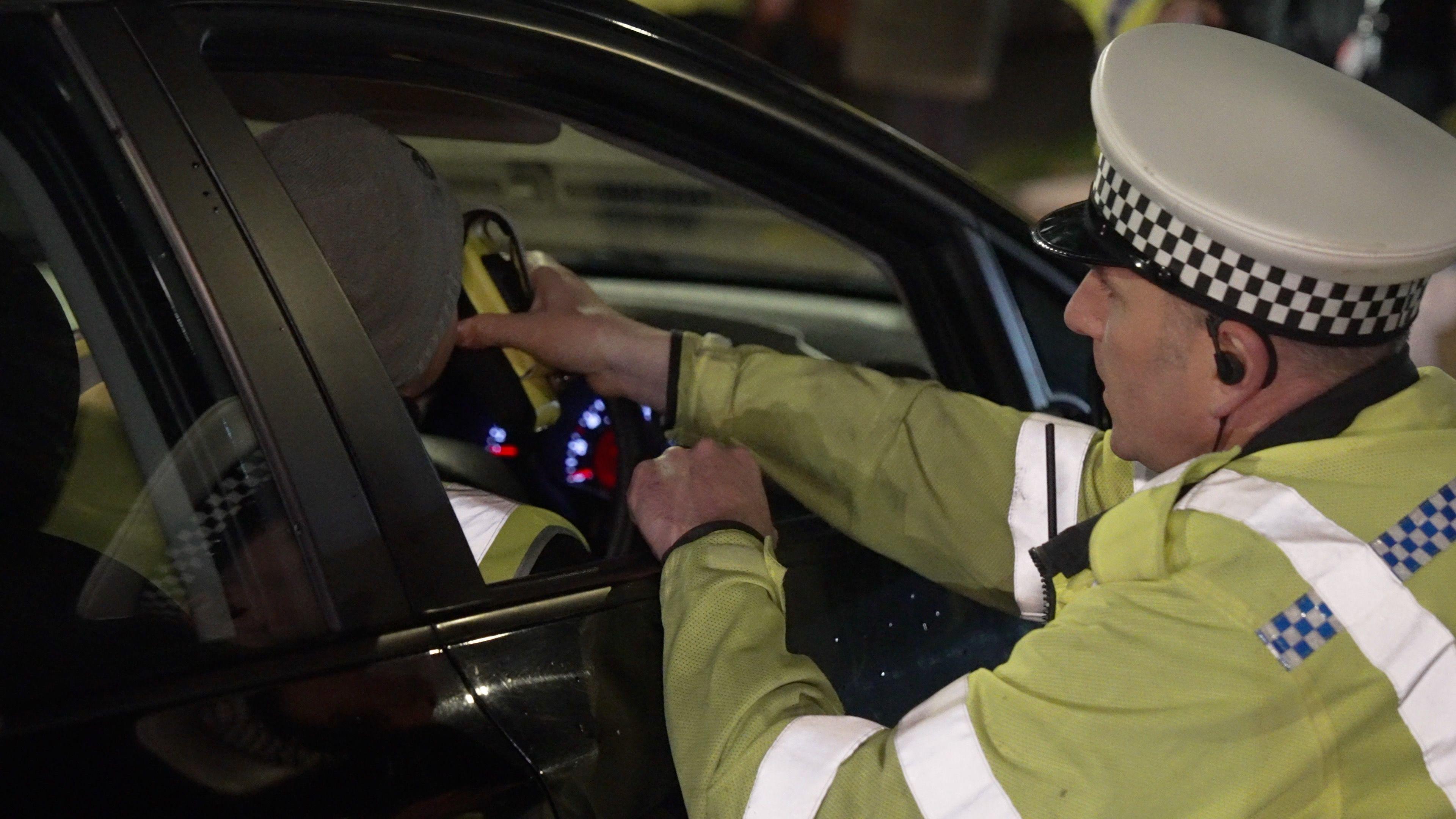 A police officer crouched down beside the driver's door of a car. He is holding a breathalyser device, which the driver of the car is blowing into. The officer is wearing a hi-vis yellow jacket and cap with black and white alternating squares around the rim.
