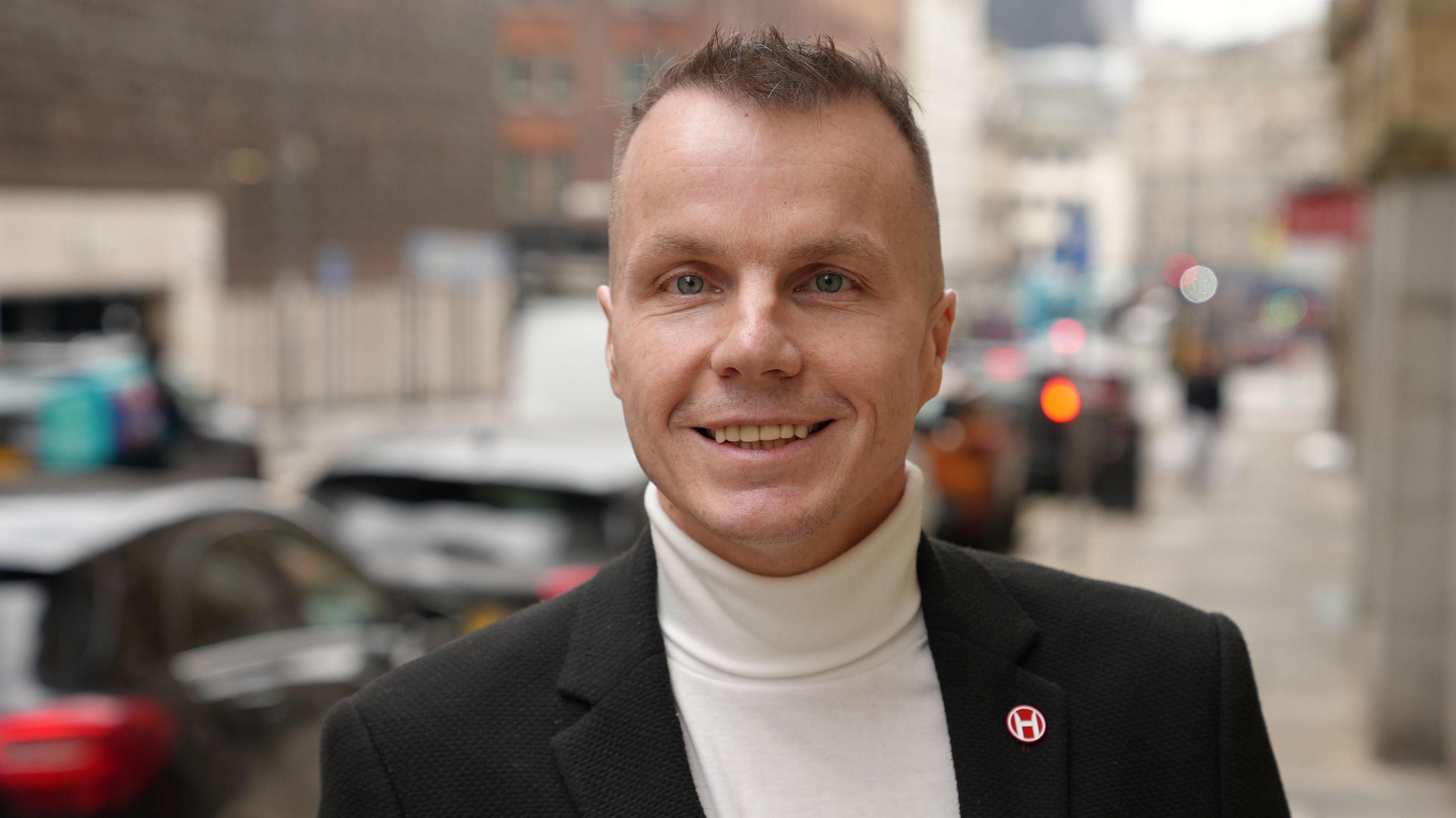 Steven Hesketh yn sefyll ar ochr stryd yn Lerpwl, yn gwisgo siaced smart ddu gyda goler uchel wen. Mae pin bach coch crwn gyda symbol gwyn wedi’i osod ar lapel chwith y siaced. Mae'r cefndir yn aneglur ac allan o ffocws, ond mae'n bosib gweld amlinelliad ceir wedi’u parcio ar hyd y ffordd ac adeiladau aml-lawr.