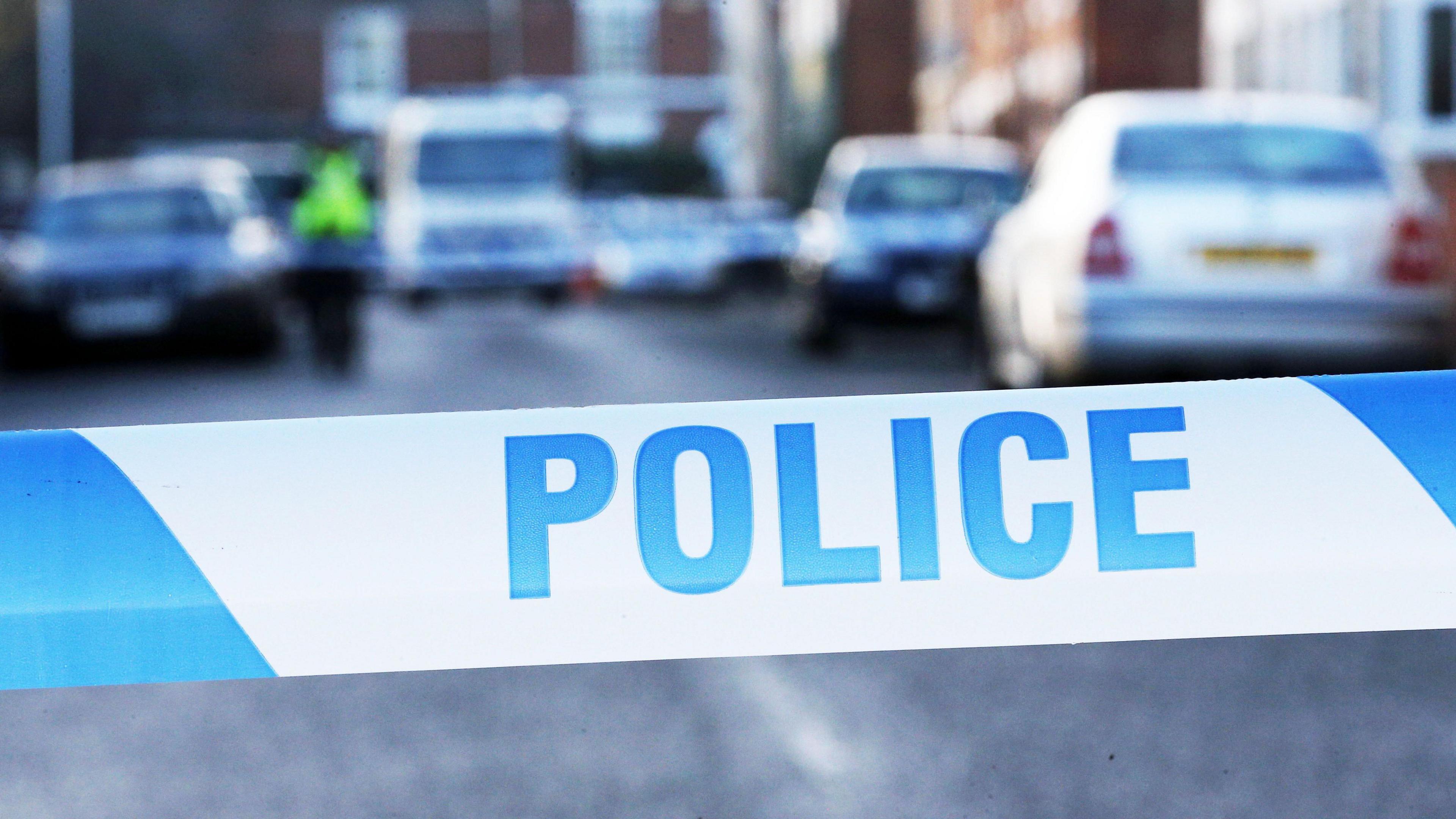 A close-up shot of blue and white police tape. It is striped with "police" written on it in blue capitals. In the background there are a number of cars on a residential street, although it is blurred quite heavily.