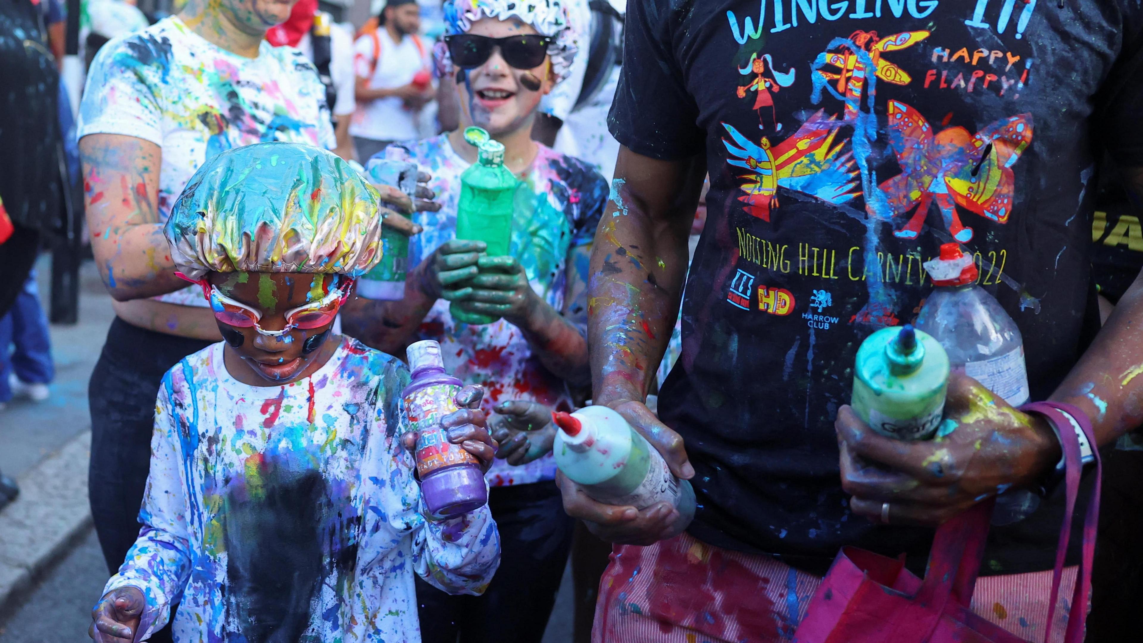 Two children in white outfits are covered in paint. One wears swimming goggles, another wears sunglasses. They carry plastic bottles of paint and are joined by two adults.