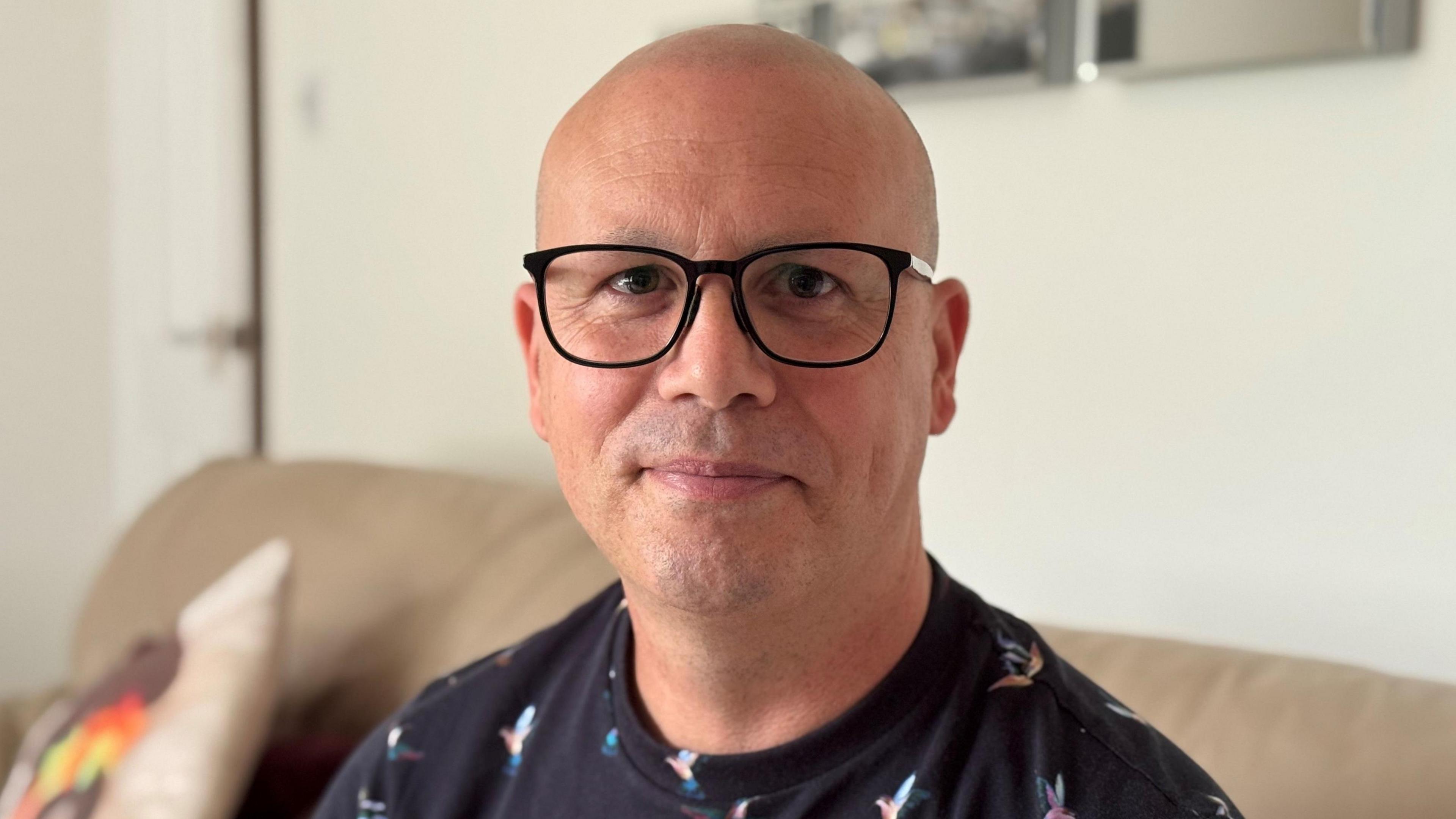 From a white-walled front room Jason Pye, with a shaven head, black-rimmed glassed and wearing a navy blue patterned top, looks into the camera lens from his beige leather sofa.