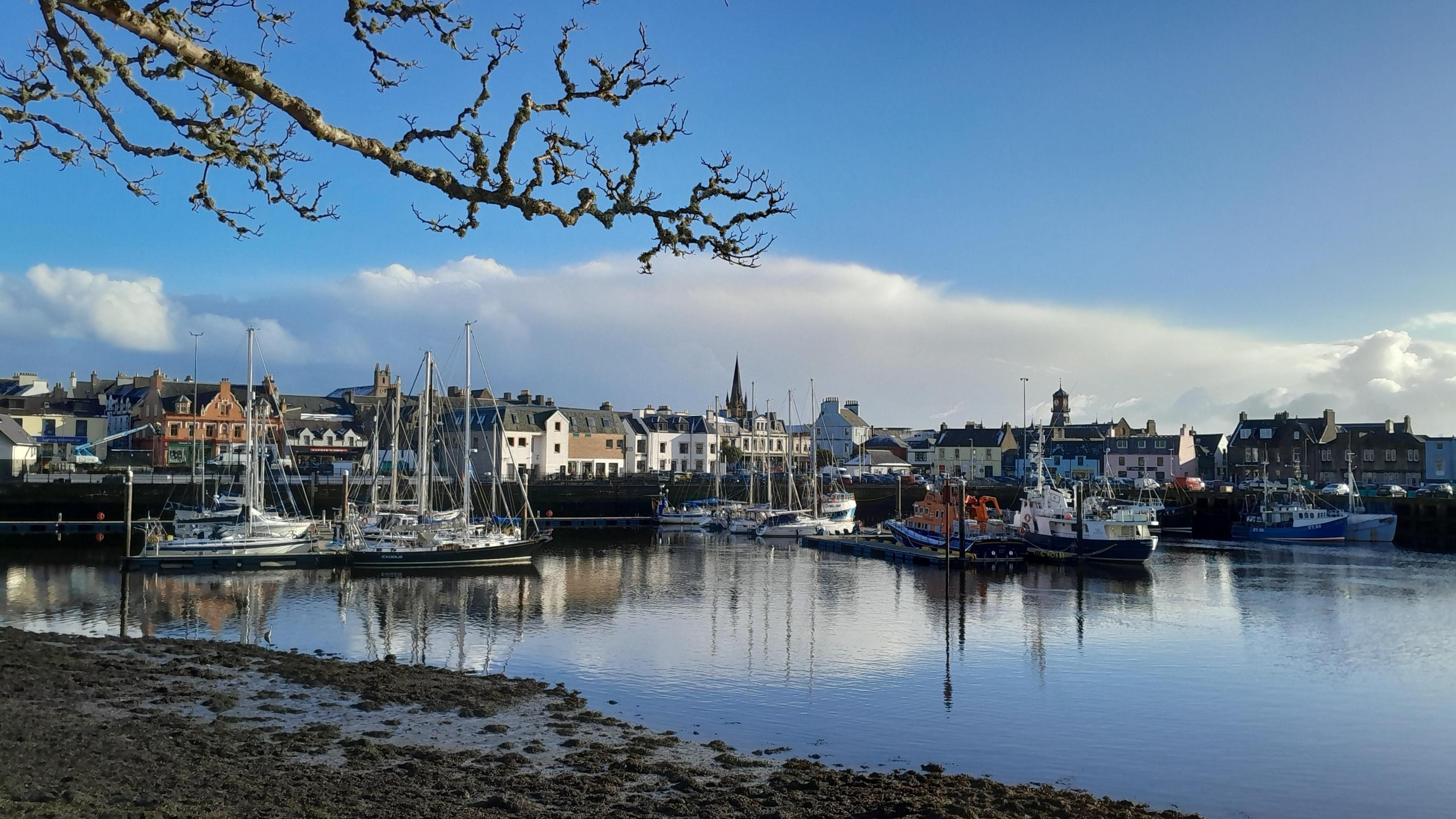 Bright blue sky with white clouds sit above a harbour with boats and lots of buildings. A muddy shore is in the foreground. 