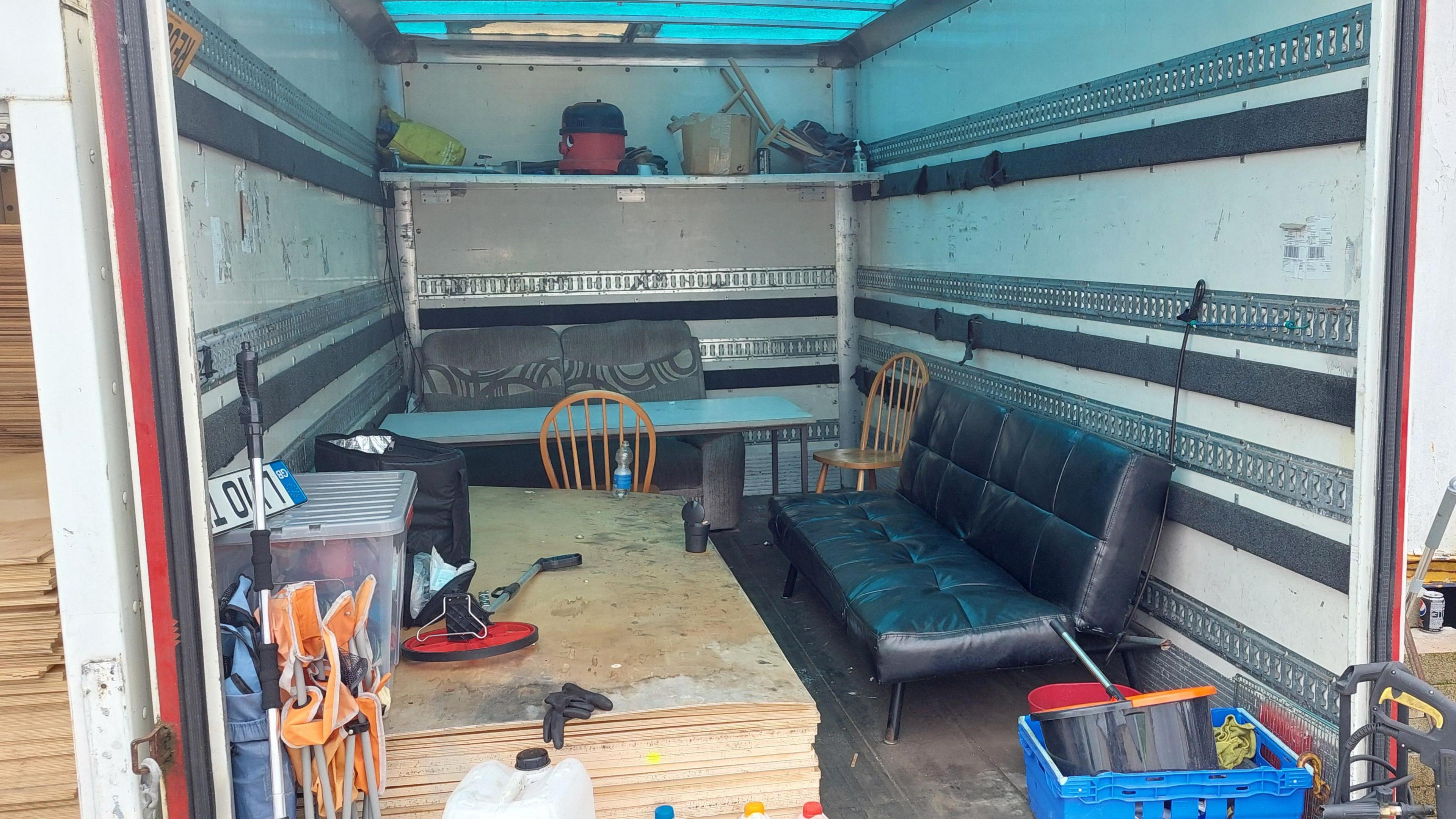 The inside of a shipping container with an old navy sofa, a pile of wooden boards, a makeshift table and car seats to sit at.