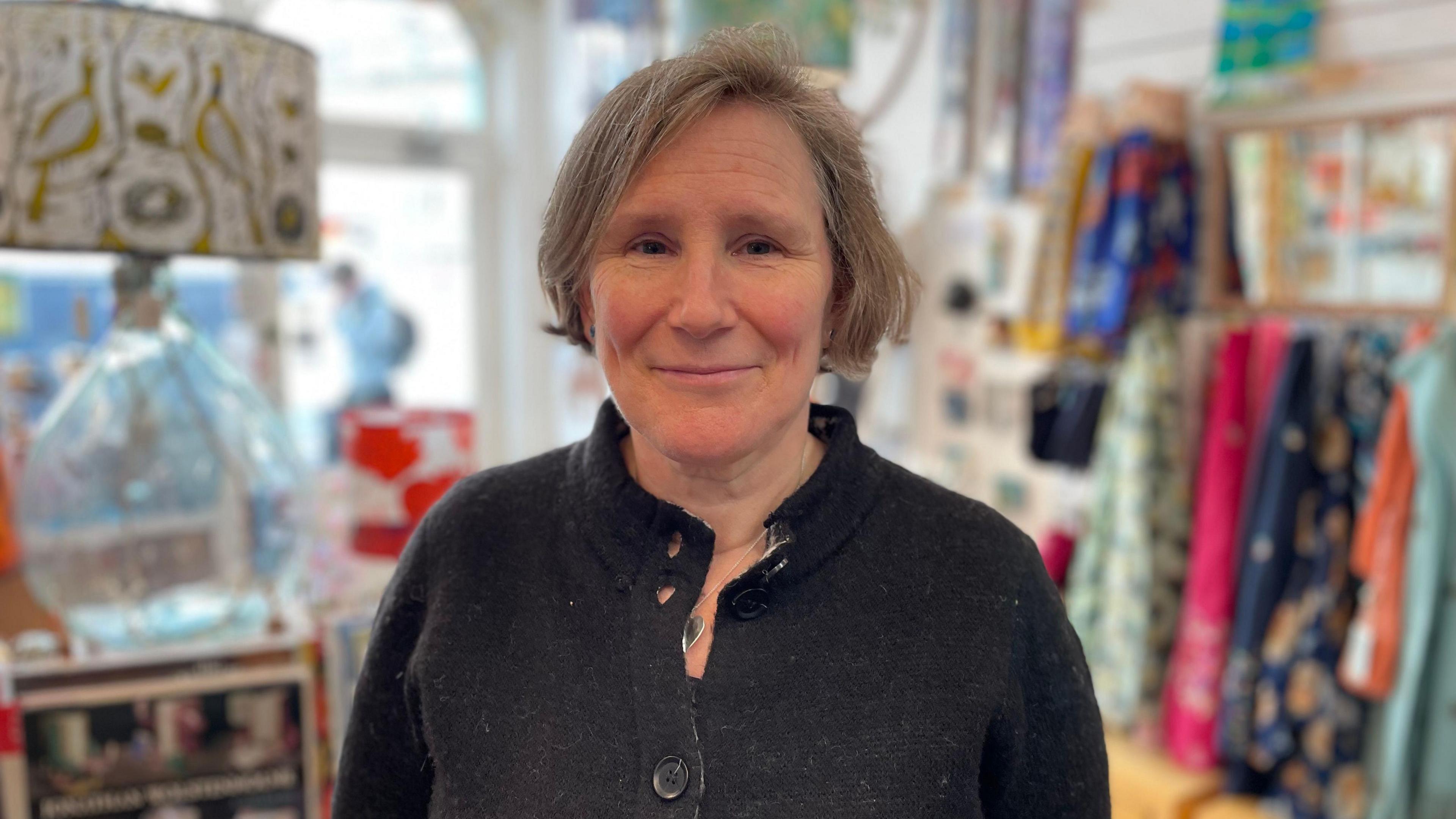 Isla Dawes standing in a shop with clothes rails, and a lamp in the background. She wears a black jacket