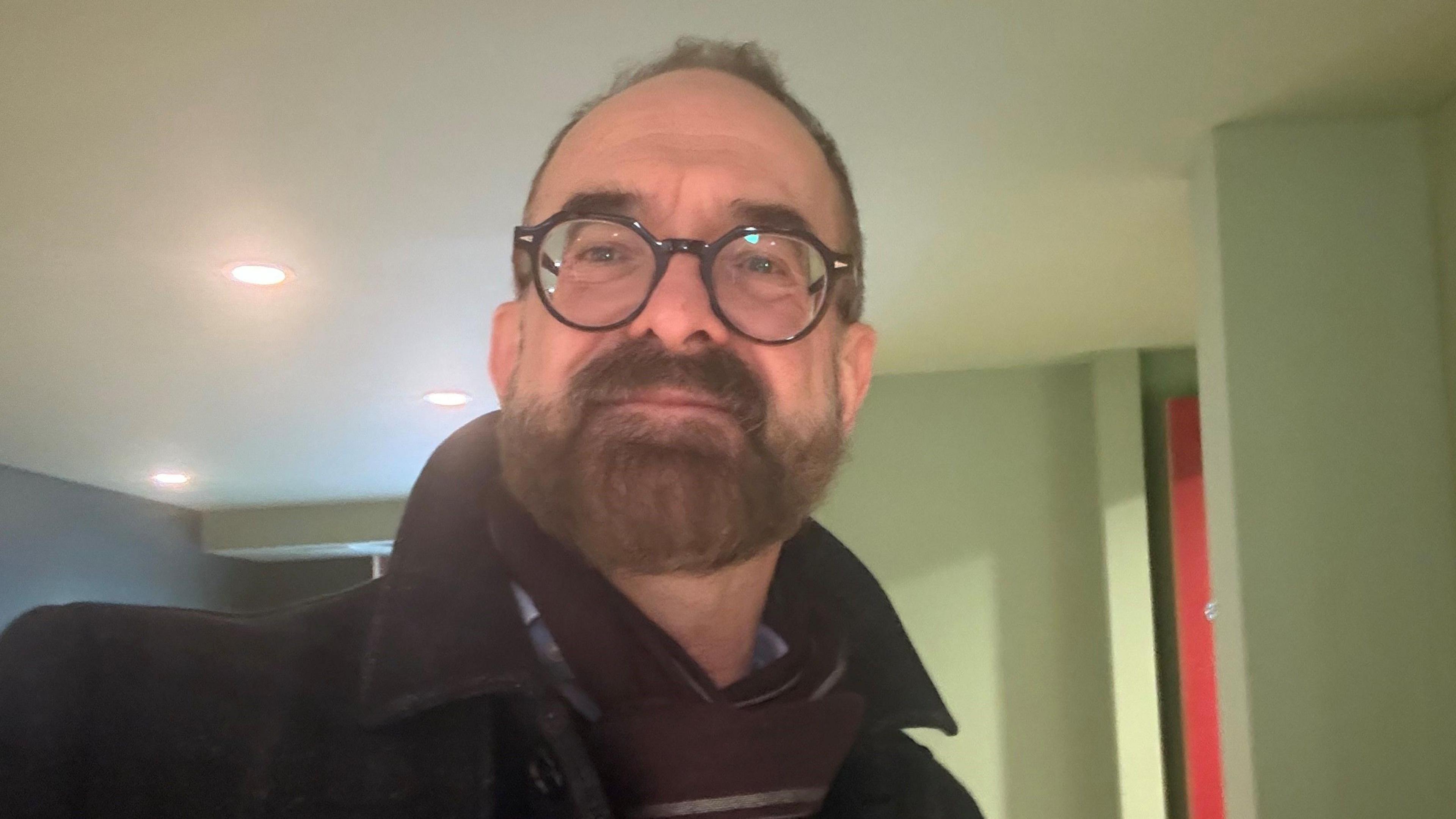 Gino Rocco, who has glasses and a beard, standing in his home with spotlights in the ceiling behind him