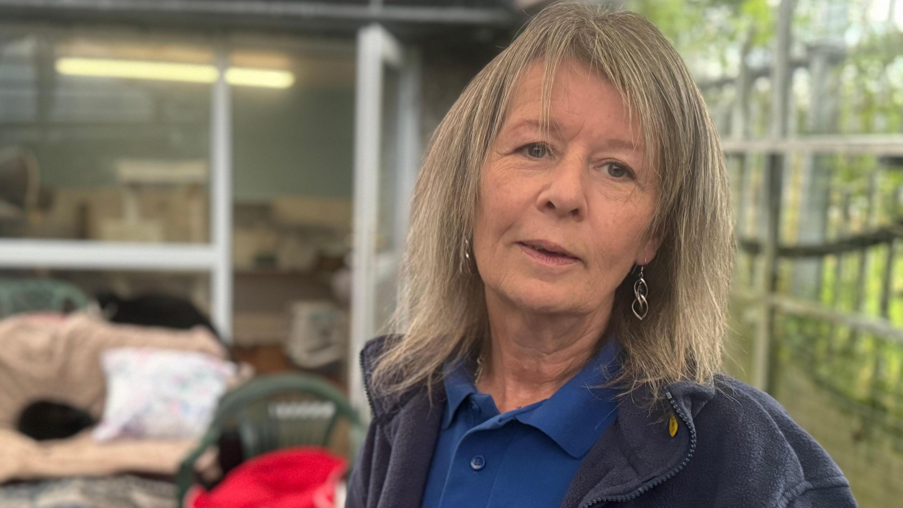 Anna looking at the camera. She has shoulder length fair hair. She is wearing silver earrings, a blue polo shirt and a navy fleece.