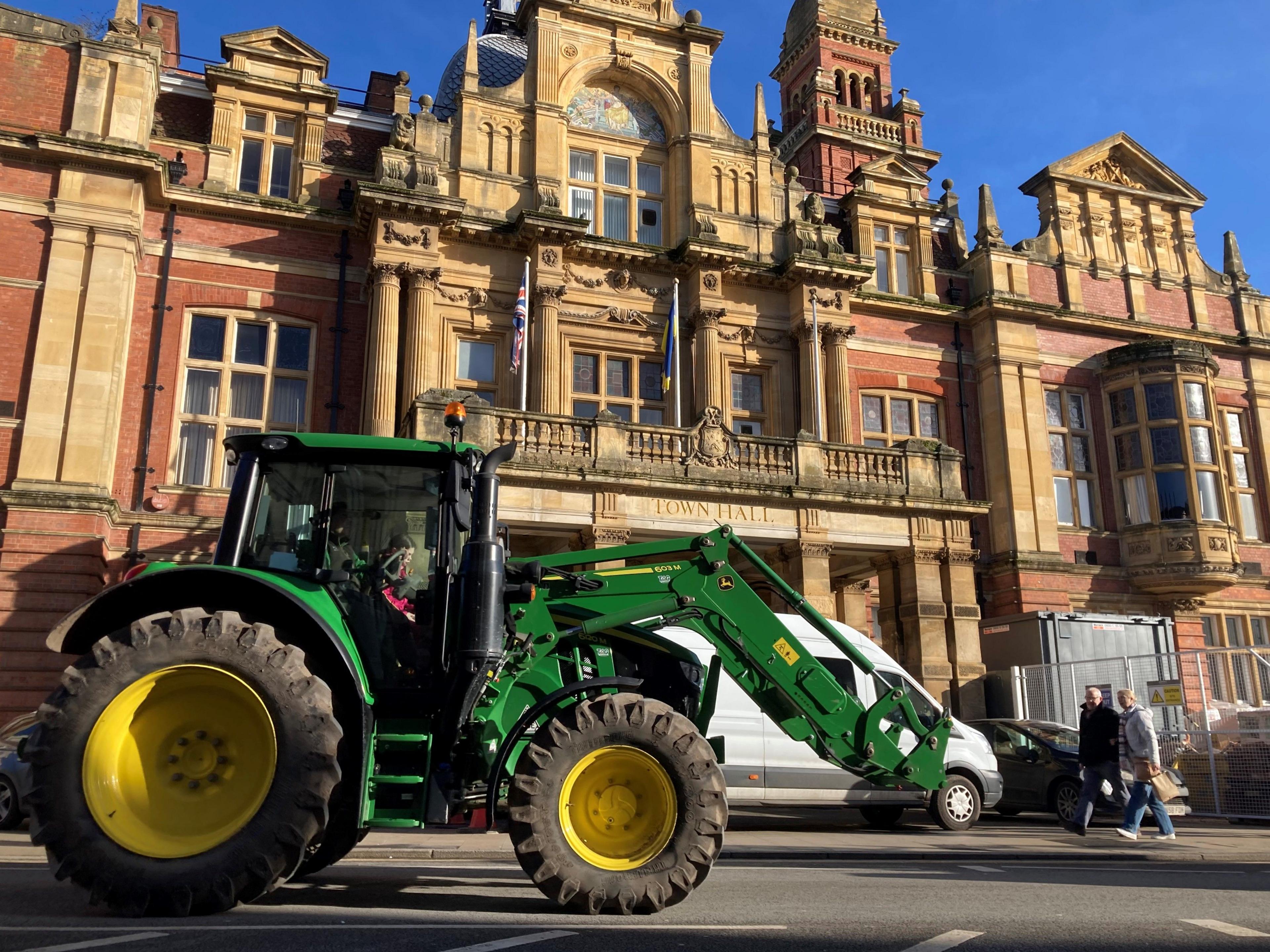 A tractor drives past a grand redbrick building on a sunny day. The tractor is green and has yellow wheels. The building has the words "TOWN HALL" engraved on the front.