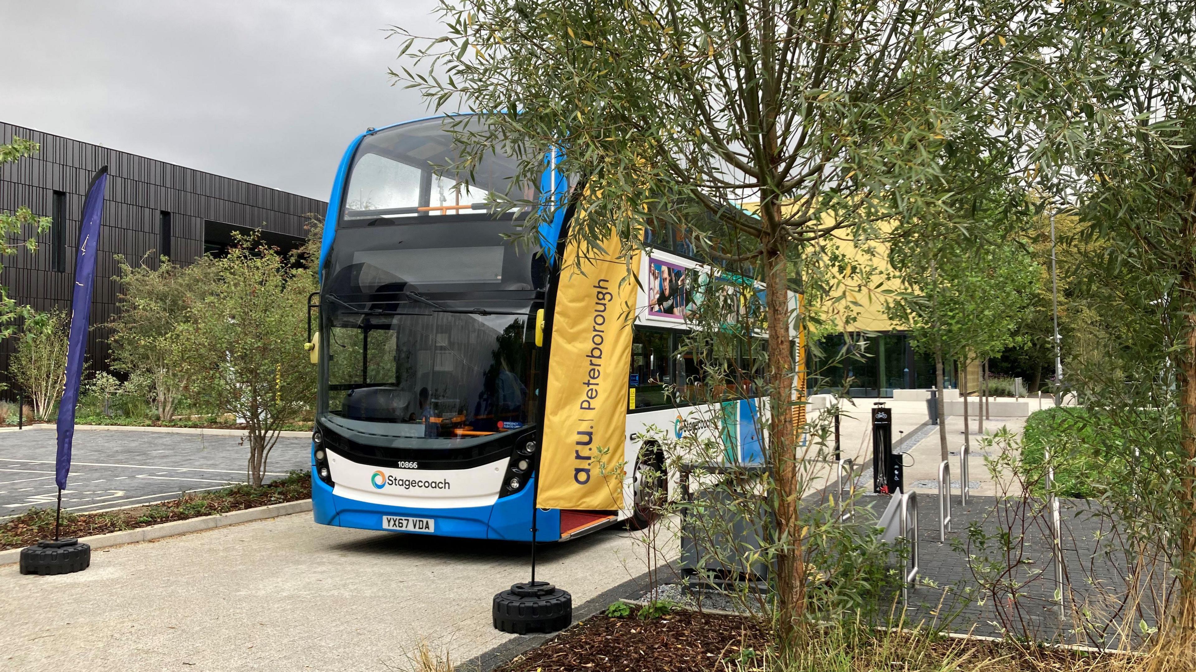 A blue and white double-decker Stagecoach bus is parked on the campus of ARU Peterborough. In front of the bus are banners with "ARU Peterborough" written on them