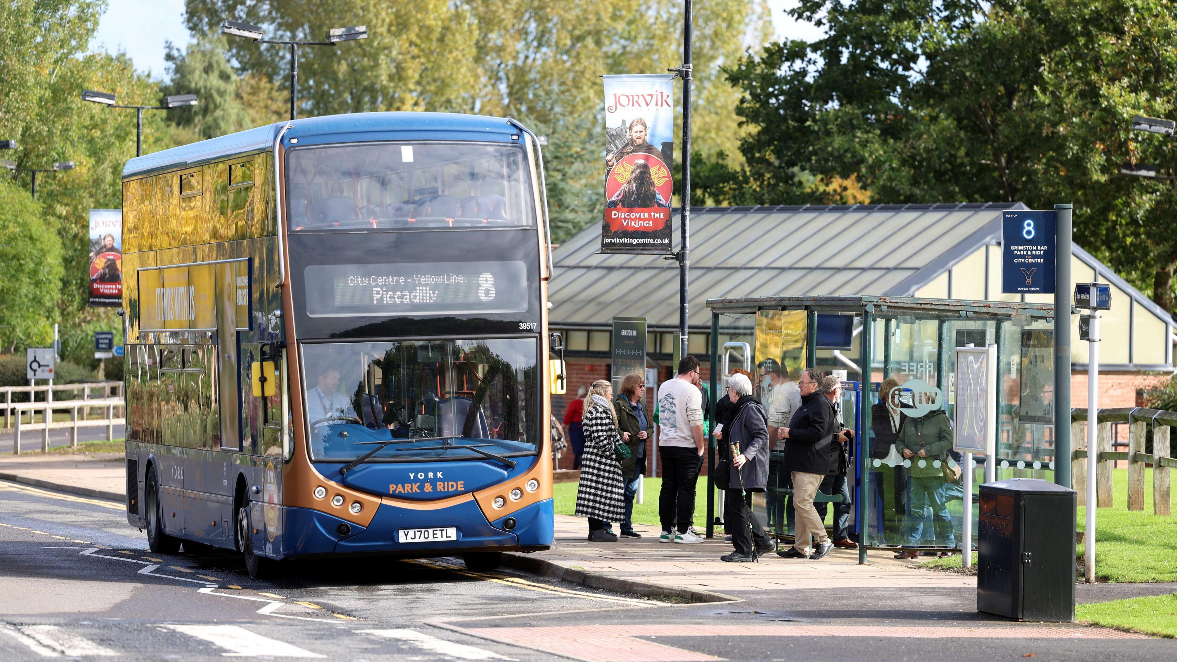 A double-decker bus marked "York Park & Ride" is parked at a sheltered bus stop. The bus displays route number 8 with the destination "Piccadilly" on its front. Several people are gathered nearby—some waiting, others boarding.