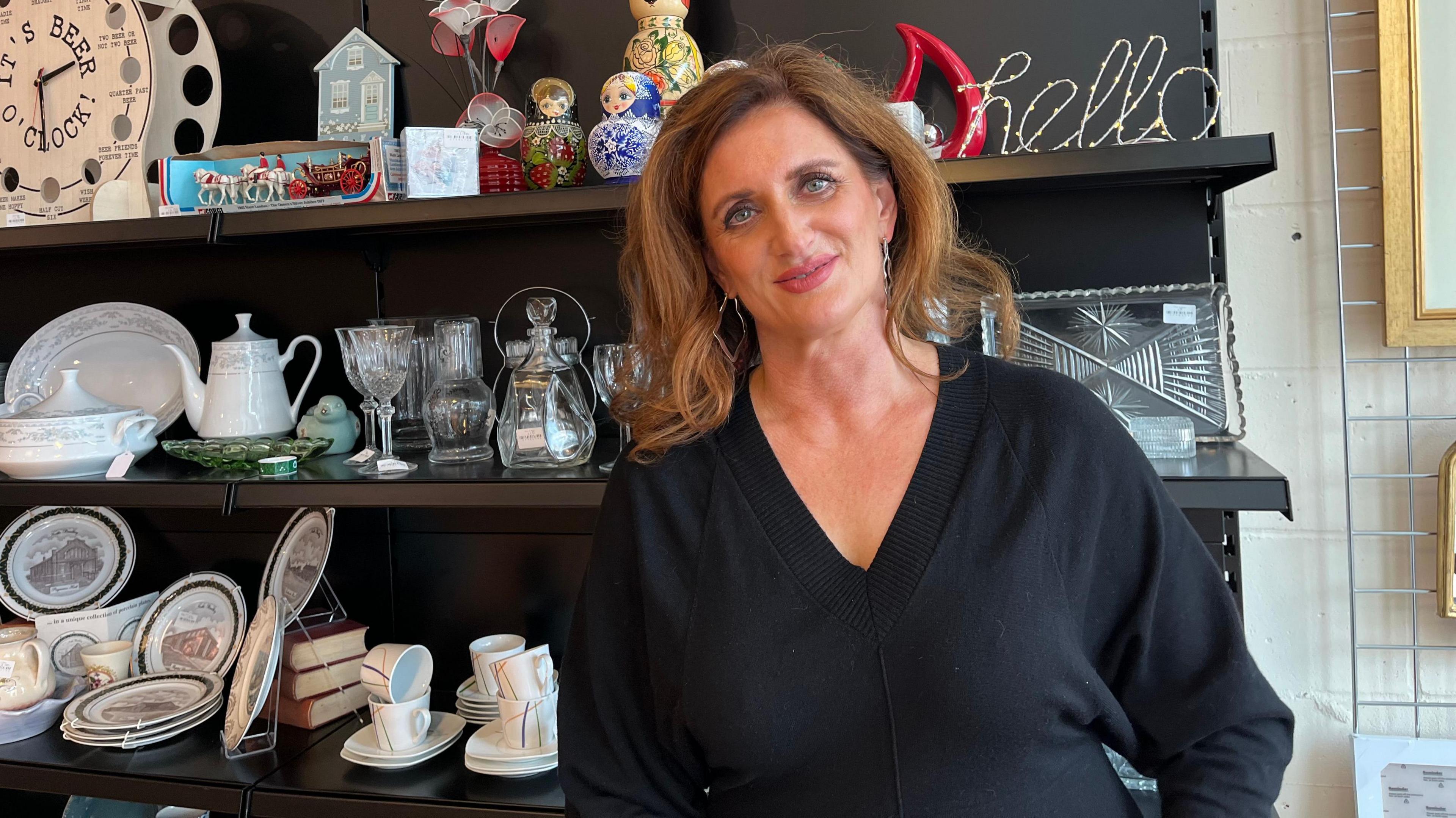 Clare Marshall is standing in front of a shelf with crockery. She is wearing a black top.