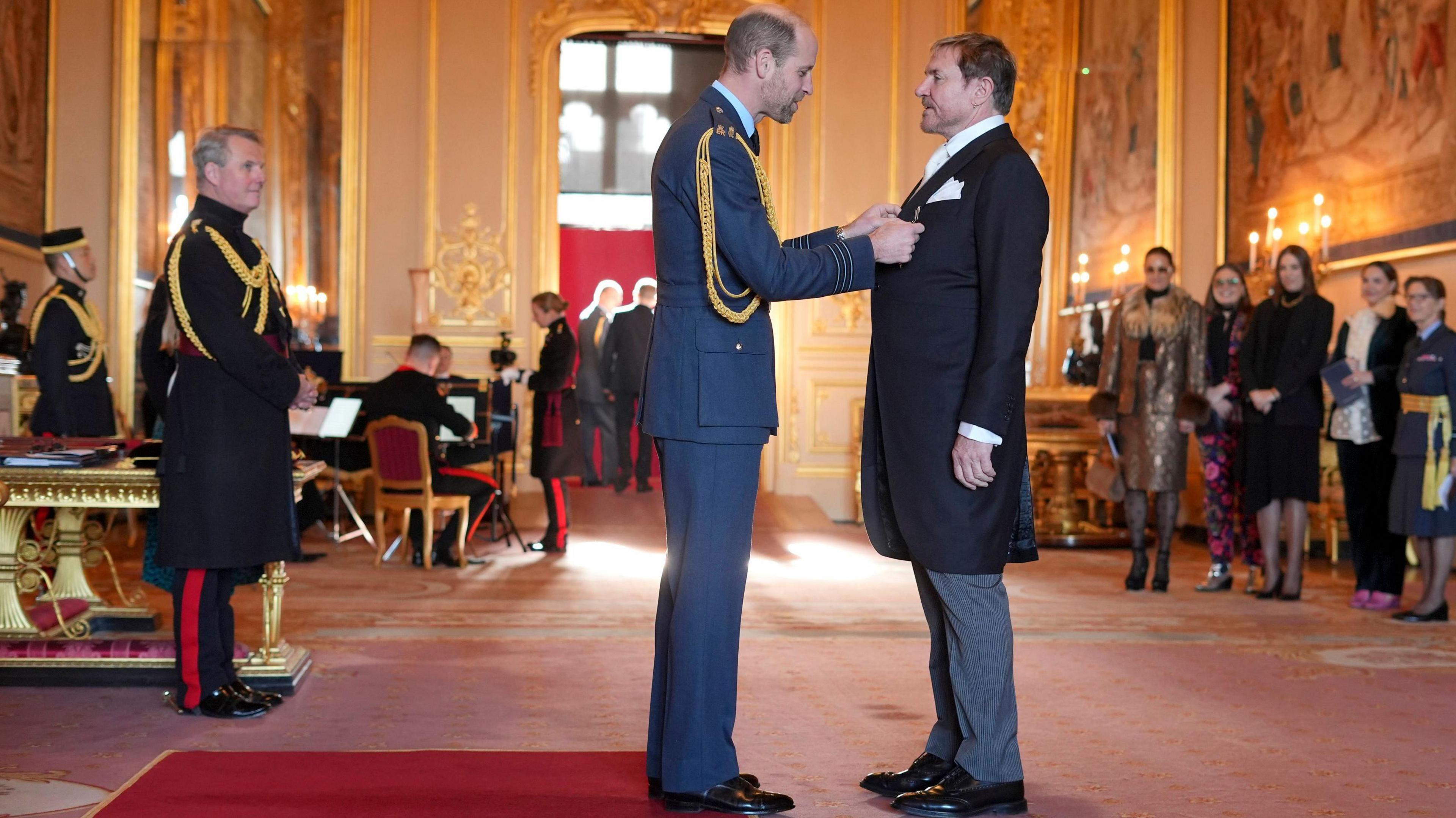 Simon Le Bon being awarded by The Prince of Wales at Windsor Castle. The room is decorated with huge paintings and red carpets. Royal staff members are present. He and Prince William seem to be engaged in a conversation. Other people could be seen behind them.