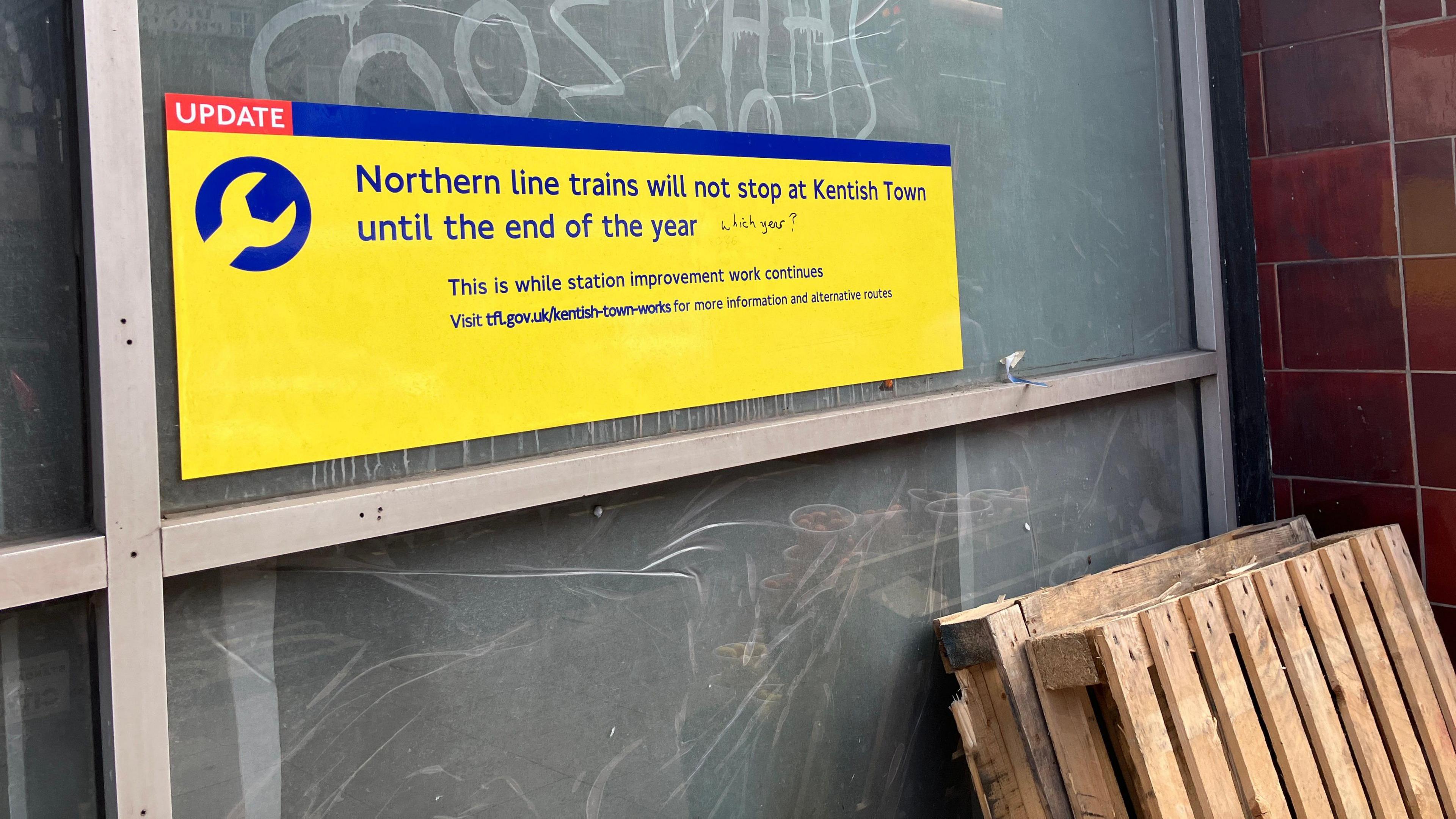 Yellow sign 'Northern line trains will not stop at Kentish Town until the end of the year' with graffiti which says 'which year?'