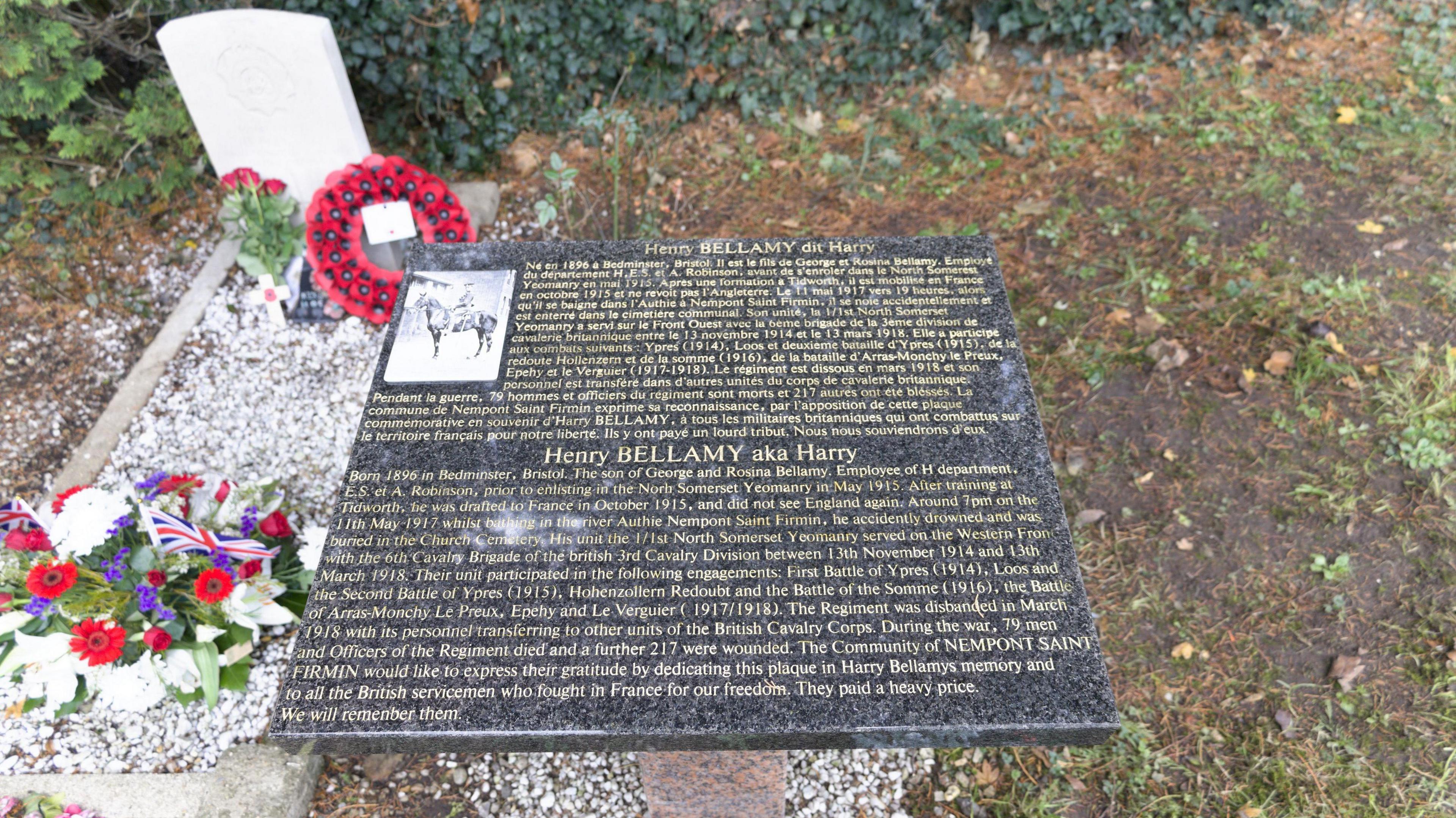 A dark marble plaque with gold writing, detailing the life of Henry Bellamy in both French and English. It details his service at the Battle of Arras in 1917 and his death. The photo of him on a horse is also on the plaque. Next to it is his grave, which has white pebbles on it, as well as flowers and a poppy wreath.